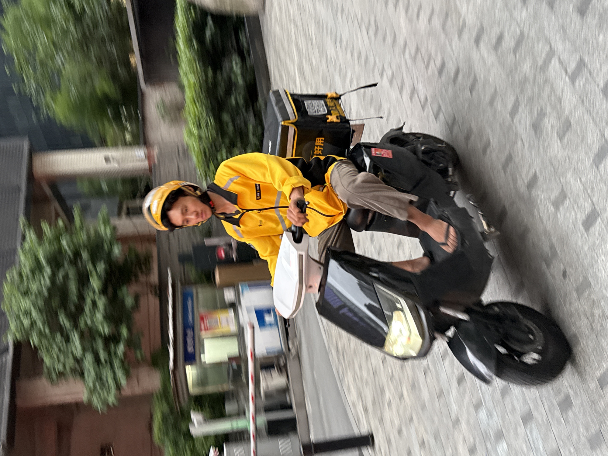 A person wearing a yellow jacket and a yellow helmet is riding a black electric scooter on a paved city street. The person is carrying a large black delivery bag on the back of the scooter. The background includes trees, buildings, and a security gate. The scene appears to be in Yuzhong District, China.