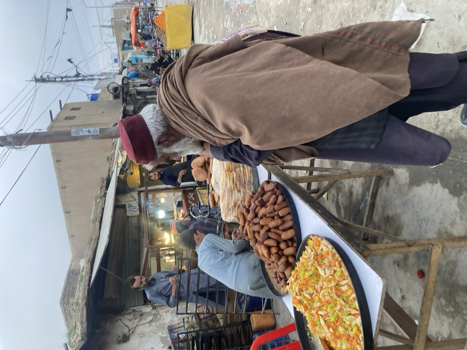 An outdoor street food market scene is depicted in Quetta City Tehsil, Pakistan, under an overcast sky. In the foreground, an elderly male with a white beard and a maroon cap, wearing a brown shawl over dark clothing, stands facing left, observing food displayed on a collapsible table. The table holds a large black tray containing fried noodles or vegetable strips, a second large black tray filled with numerous elongated brown fried pastries or fritters, and a tall stack of round flatbreads. A younger male in a light blue garment is positioned behind the table, leaning over the food. In the midground, another bearded male in a light blue shalwar kameez stands behind the food stall. Further left, a third bearded male in a dark top is visible inside an adjacent open-front shop, which appears to be a bakery with metal racks and illuminated display cases. The street surface is concrete and appears damp in areas. A prominent utility pole with numerous overhead wires occupies the right midground. In the background, low-rise buildings with weathered facades line the street, extending into a busy market area with additional stalls, including one featuring oranges in a yellow cart.