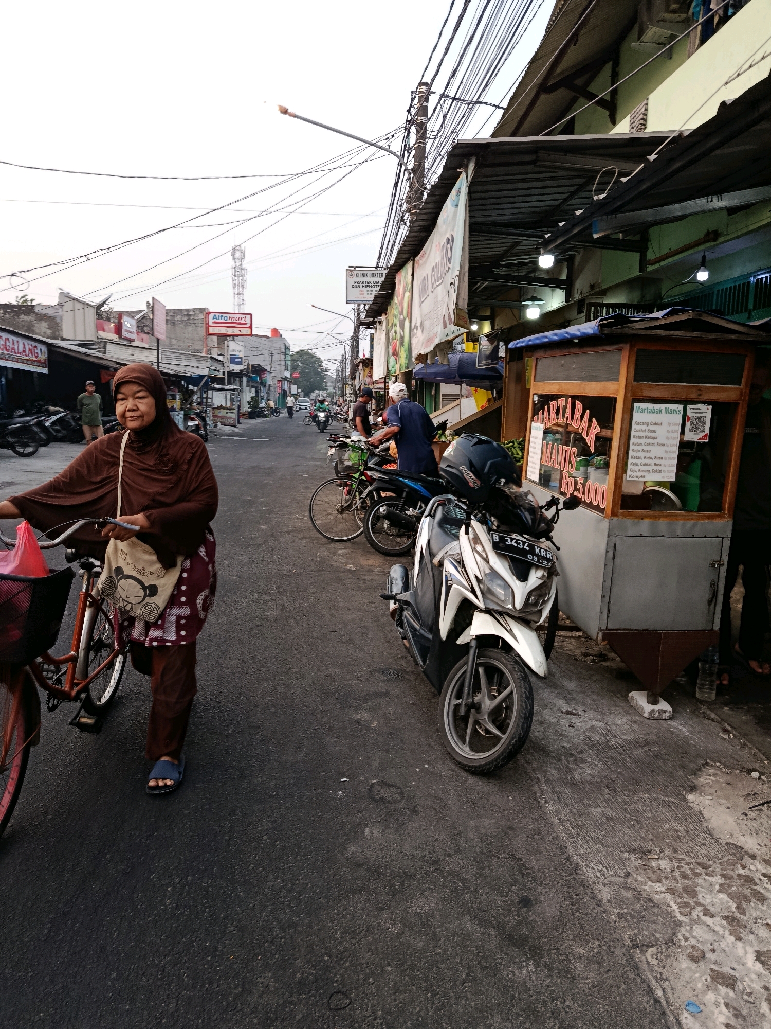 The image depicts a street scene in Bekasi, Indonesia, likely during the early morning or late afternoon, given the soft lighting and overcast sky. In the foreground, a woman wearing a brown hijab and brown attire is pushing a bicycle. She has a bag slung over her shoulder with a cartoon-like drawing on it. The bicycle has a basket in the front, partially filled with items. Behind her, a white and black scooter is parked.
To the right of the scooter is a food cart selling "Martabak Manis," indicated by signs on the cart. The menu lists various toppings such as chocolate, peanuts, and cheese, with prices starting at Rp5,000. The cart itself is made of wood and has glass panels.
Further down the street, various buildings line both sides, with awnings providing shade. A sign for "Alfamart," a convenience store chain common in Indonesia, is visible. Other signs for businesses and advertisements are also present. Several motorcycles are parked along the side of the road, and a few people are walking or standing. The road surface appears to be asphalt, with some patches of wear and tear. Power lines crisscross overhead. In the distance, a cellular tower can be seen. The overall impression is that of a bustling local street with commercial activity.