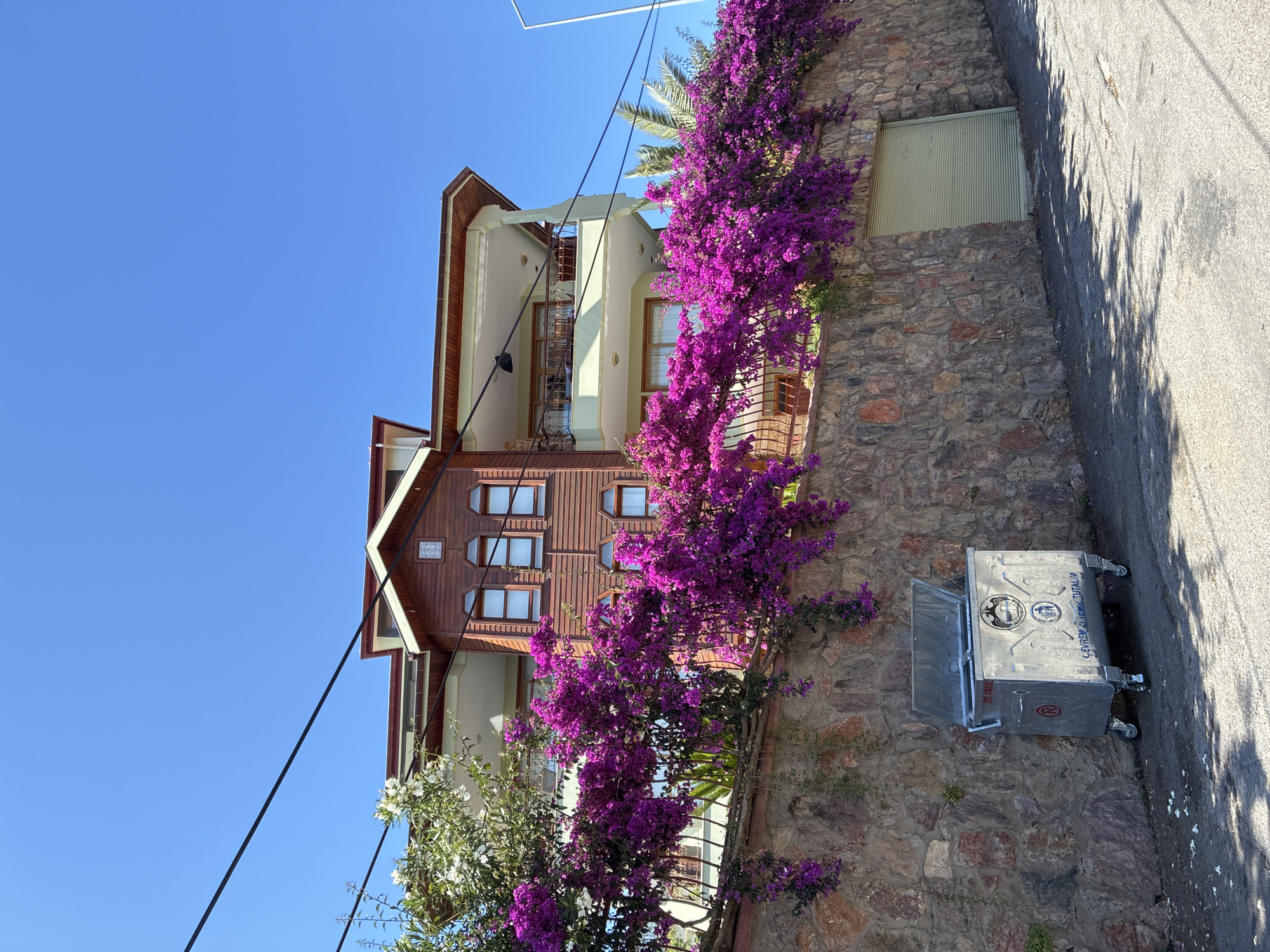 This outdoor image depicts a sunny residential scene in Alanya, Turkey, characterized by a clear blue sky. A multi-story building stands behind a substantial stone wall, with its upper section featuring a brown wooden facade, multiple arched windows, and a reddish-brown tiled roof. The lower visible part of the building has light green walls and balconies with decorative metal railings.
The stone wall, constructed from varying shades of brown, red, and grey stones, runs along an inclined asphalt road. Abundant vibrant purple bougainvillea cascades over a metal fence atop the stone wall, with some white flowering plants visible on the left side near the building. Towards the right, a light green corrugated metal door is embedded within the stone wall.
On the left side of the asphalt road, a large, wheeled, metallic refuse bin with a partially open lid is positioned against the stone wall. The bin is adorned with Turkish text, including "ÇEVREMİZİ TEMİZ TUTALIM" (Let's keep our environment clean) and "ATEŞ ATMAYINIZ" (Do not throw fire), along with a municipal emblem and waste disposal symbols. Black utility wires diagonally traverse the upper portion of the sky, and a white street light pole is partially visible on the far right. Shadows from unseen objects are cast across the road surface. No individuals are present in the image.