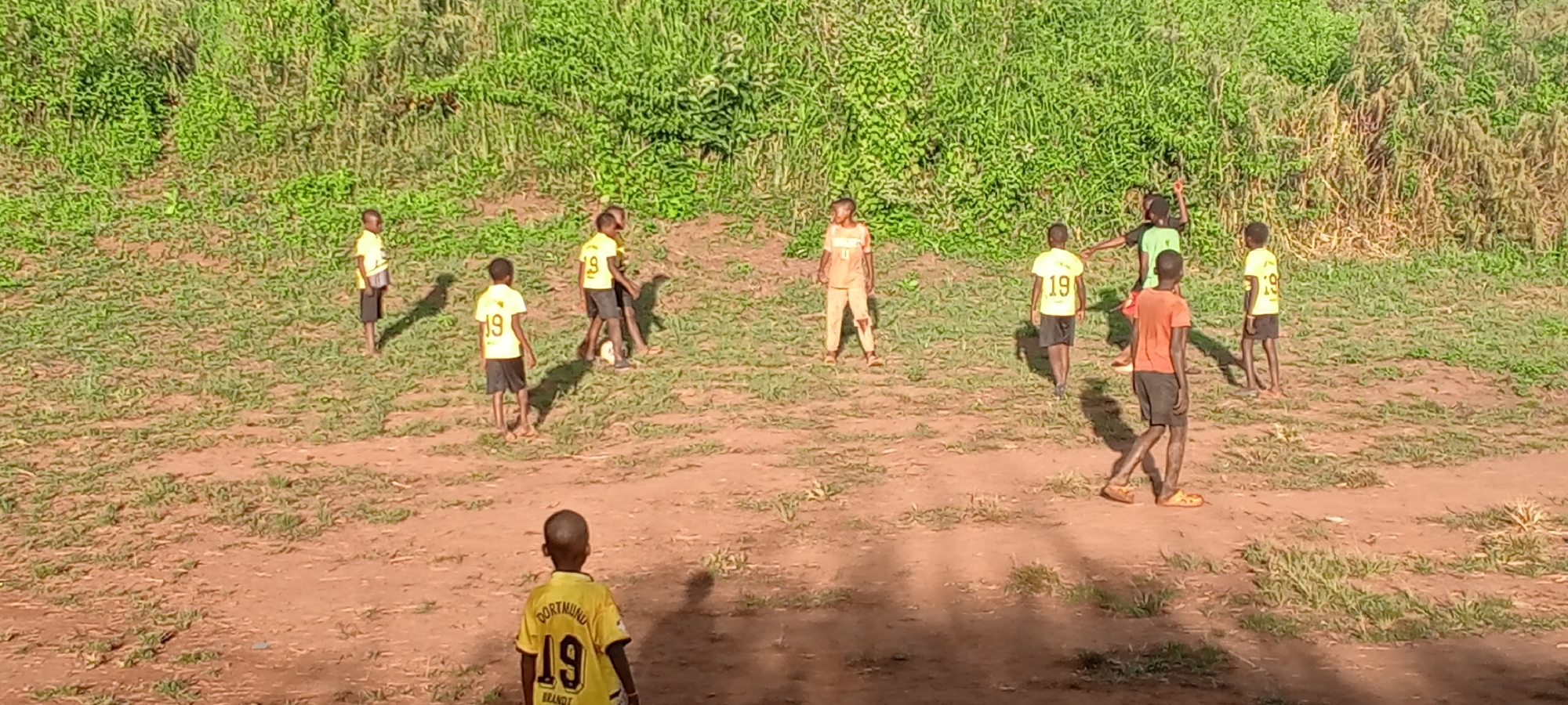 A group of children are playing soccer in Ngalula, Democratic Republic of the Congo. Nine boys are visible; at least three wear yellow soccer jerseys with the number "19" on the back. One child wears an orange shirt, and another wears green. The boys are in a grassy area, partially dirt, with a dense green backdrop of vegetation, possibly a hillside. Some of the boys are interacting around a soccer ball, while others are standing apart, possibly waiting for their turn. The time of day appears to be sunny, indicated by shadows on the ground.