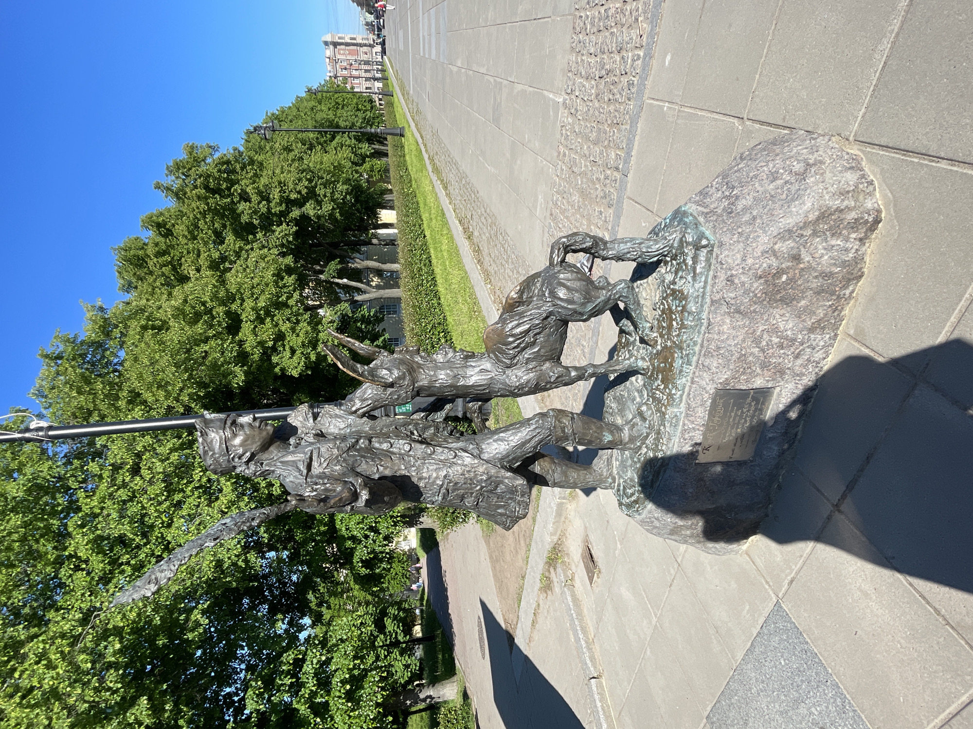 The image displays a bronze sculpture situated outdoors on a clear, sunny day in Saint Petersburg, Russia. The sculpture depicts a dynamic scene featuring a person, likely a hunter or explorer, in historical attire with a large hat, holding a long staff or spear. They are shown in confrontation with a wild boar, characterized by its prominent tusks, which appears to be lunging or in motion. The figures exhibit textured detailing, typical of bronze artwork, and the sculpture is mounted on a large, rough-hewn stone base.
In the background, a wide, paved pedestrian path or avenue stretches into the distance, flanked on one side by neatly trimmed green hedges and lush deciduous trees. Beyond the trees, buildings with classic urban architecture are visible under a bright, cloudless blue sky. The strong shadows cast on the pavement indicate either morning or afternoon sunlight.
A metal plaque is affixed to the stone base of the sculpture. Partial Cyrillic text is visible on the plaque, with the first line beginning with "К", the second line possibly starting with "В", and a subsequent line starting with "Ф" or similar letters, providing information or a dedication related to the artwork. The overall setting suggests a public park or a significant city boulevard.