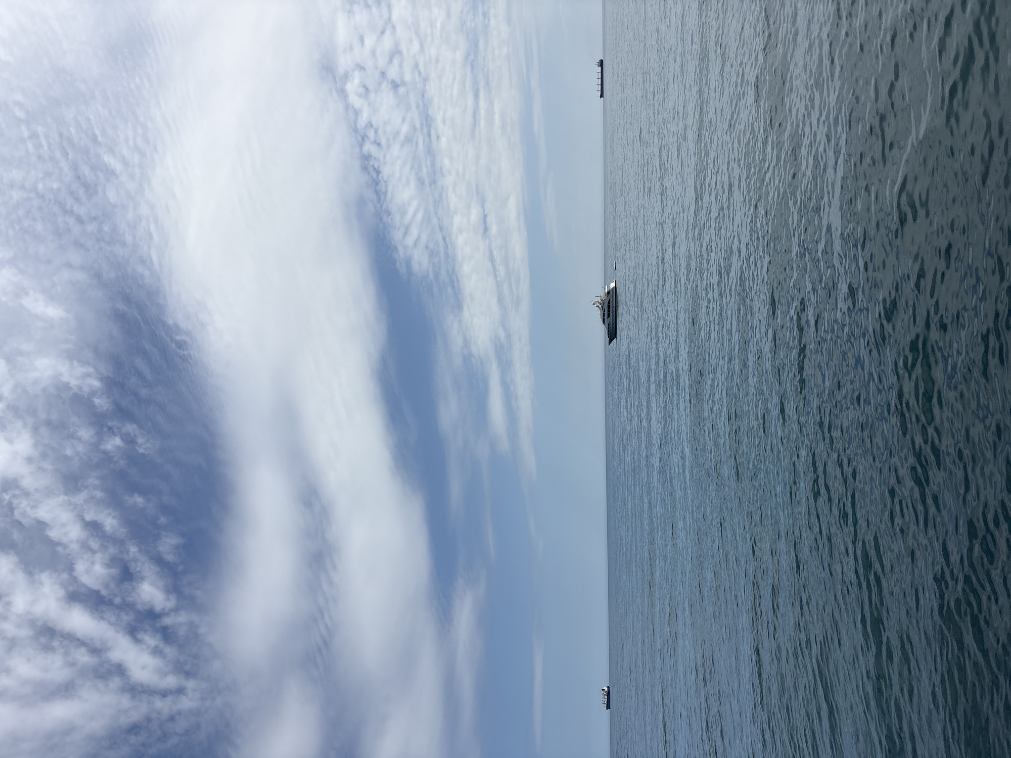 The image depicts a view of the sea and sky in Ялта, Ukraine. The right side of the image shows a body of water with visible texture indicating slight waves. Two vessels, possibly ships or boats, are present in the water, one closer to the center and the other in the distance towards the top. The left side of the image showcases a sky with patches of blue and clouds. The overall composition suggests a serene, open environment with a coastal perspective.