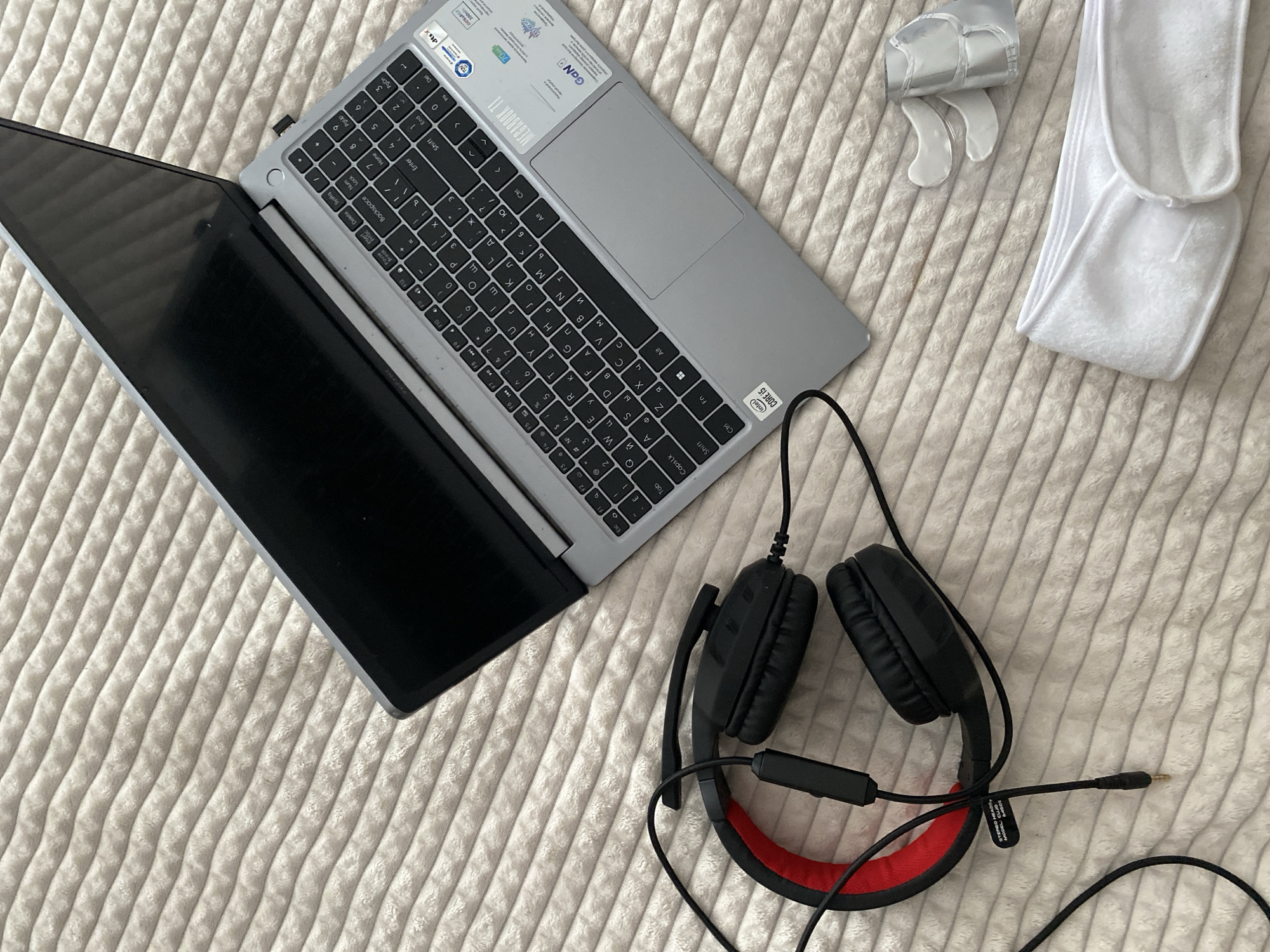 The image shows a laptop computer, headphones, and cosmetic accessories on a textured, light-colored surface, possibly a bedspread or blanket. The laptop is open, displaying a dark screen and a keyboard with Cyrillic characters. The headphones are black with red accents. Next to the laptop are cosmetic accessories: white headbands and eye patches. There is nothing to indicate the city or location in Sochi, Russia.