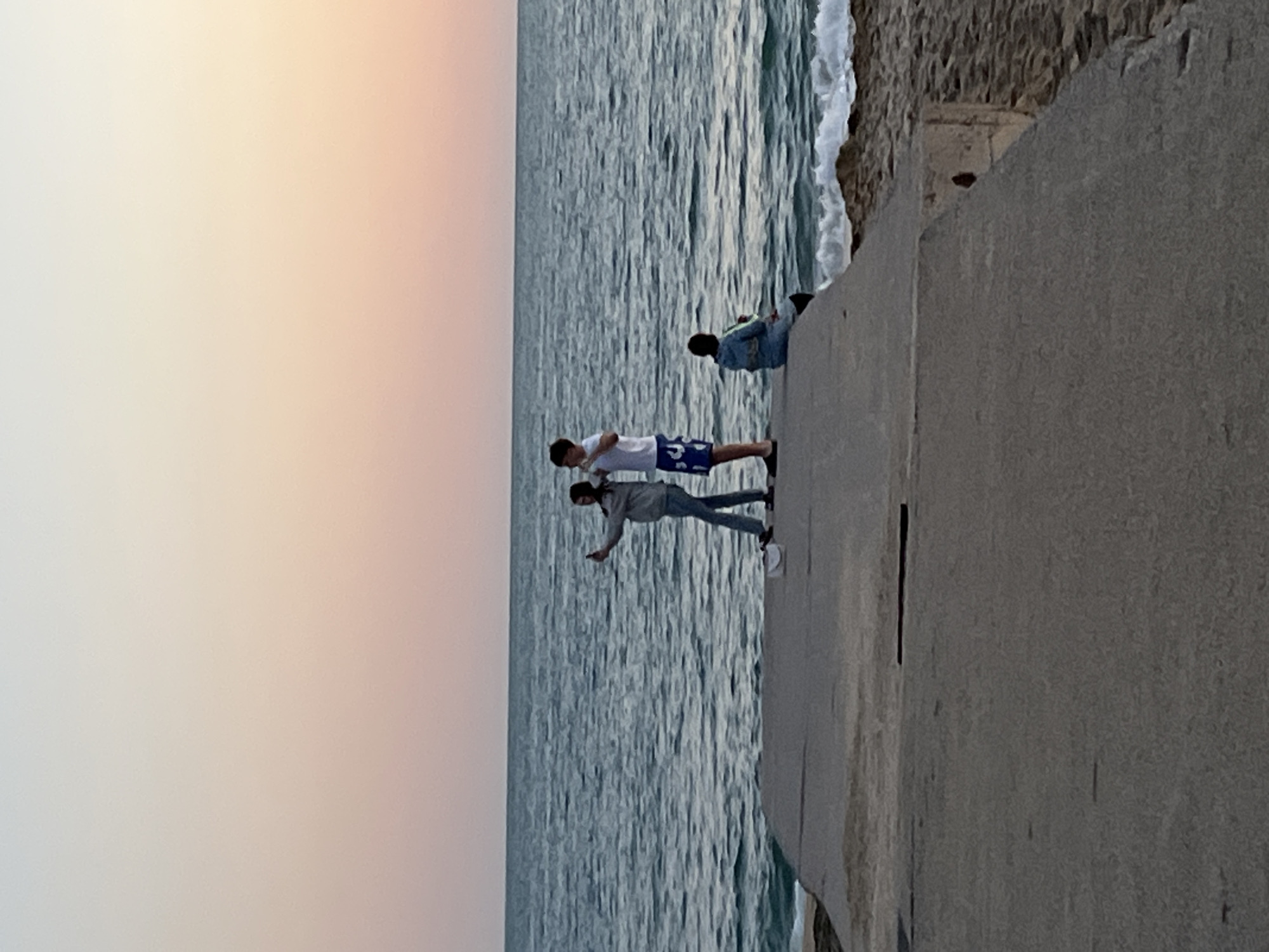The image shows a coastal scene, likely at sunset, featuring a concrete pier with individuals standing on it. Two young males are standing close to the edge, one with his arm outstretched, possibly pointing. A third person is seated on the pier further back. The sea is visible on the left side. The sky is visible to the left, appearing gradient, ranging from light to darker shades, indicating either sunrise or sunset. The picture was taken on Alanya beach in Alanya, Turkey.