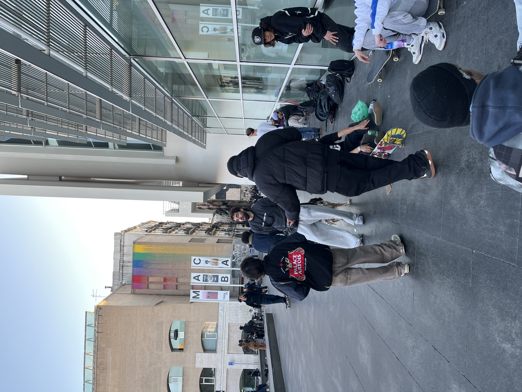 The image shows a group of young people, many with skateboards, gathered outside the MACBA (Museu d'Art Contemporani de Barcelona) in Barcelona, Spain. Some individuals are sitting or lying on the ground, some are standing, and others appear to be in conversation or resting. Several skateboards and backpacks are scattered on the ground. The architectural details of the museum and surrounding buildings are visible, including the modern glass facade of the MACBA, the older brick buildings across the plaza and a colorful mural on one of the buildings. The weather appears to be clear and sunny.