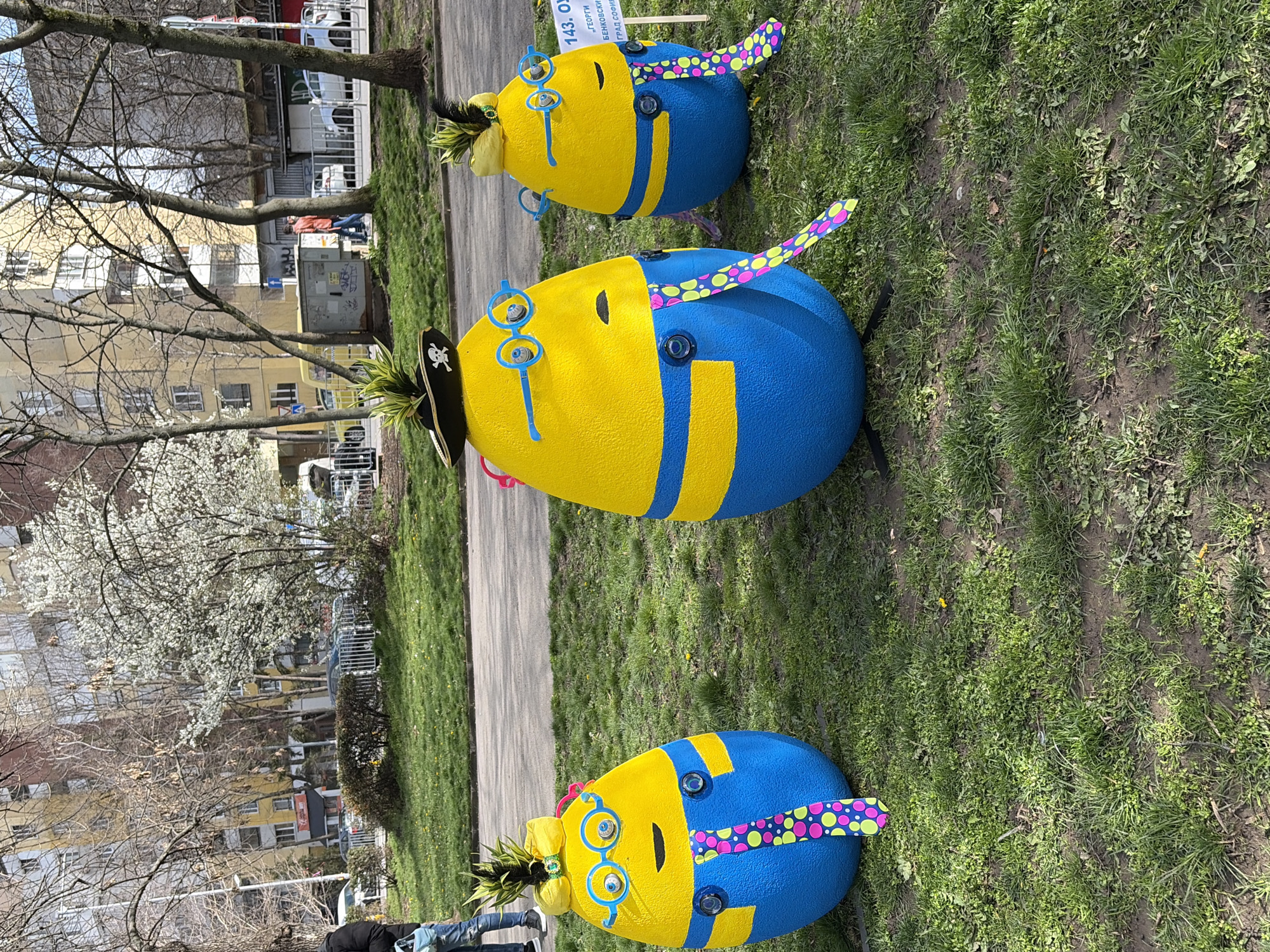 The image shows a row of three egg-shaped, painted figures in a grassy area near a sidewalk or street in Sofia, Bulgaria. The figures are painted yellow and blue, resembling Minions. Each has a tuft of green foliage on top, resembling hair. Two of the figures wear blue glasses and have polka-dotted neckties. The middle figure also wears a black pirate hat. Buildings and trees are visible in the background, with some trees in bloom. A sign with Cyrillic text is partially visible, possibly indicating a school or organization. The scene is outdoors during the day.