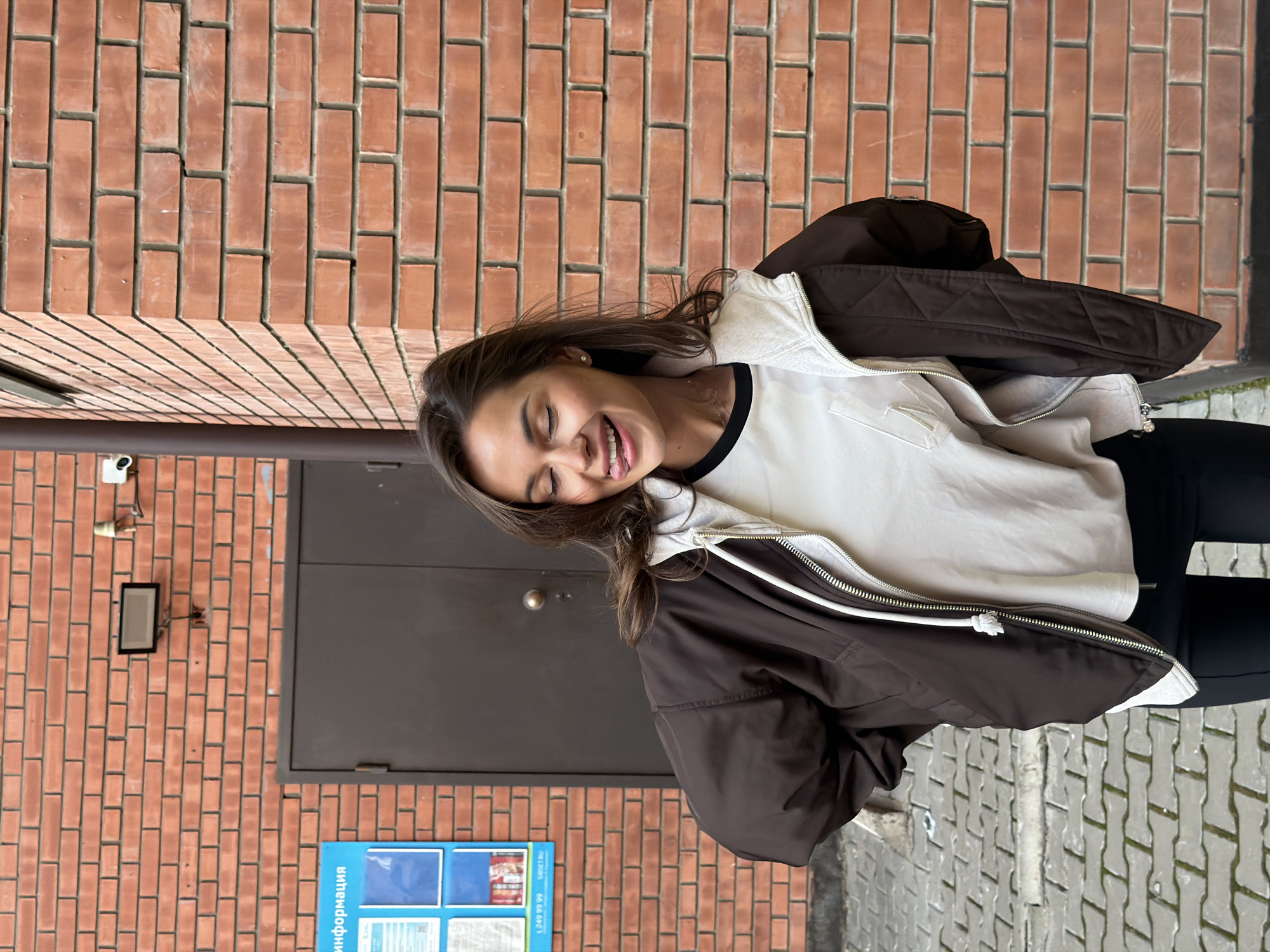 A vibrant, horizontally-oriented outdoor portrait captures a young woman laughing heartily against a textured brick wall. She is positioned to the right of the frame, her eyes closed in mirth, and her mouth open with her tongue slightly visible, conveying genuine joy and playfulness. Her dark hair flows casually as she leans her head back slightly.
She is dressed in a dark brown puffer jacket, worn open over a light-colored, possibly off-white, long-sleeved top with a black-rimmed collar. Black leggings or pants complete her attire.
The setting is an urban outdoor space, likely a street or walkway beside a building in Krasnoyarsk, Russia. The background features a reddish-brown brick wall with dark mortar lines. On the wall, to the left, a dark gray door or panel is visible. Above it, a white security camera and a black rectangular outdoor light fixture are mounted. A vertical metal downspout runs along the left edge.
The ground is paved with interlocking gray blocks, with some moss or dirt visible in the crevices. In the lower left corner, a blue information stand is partially visible. The text "информация" (Russian for "information") is clearly legible, along with a partial phone number "249 99 99". The scene is illuminated by bright daylight, suggesting fair weather. The overall mood is cheerful and candid, capturing a spontaneous moment of happiness.
