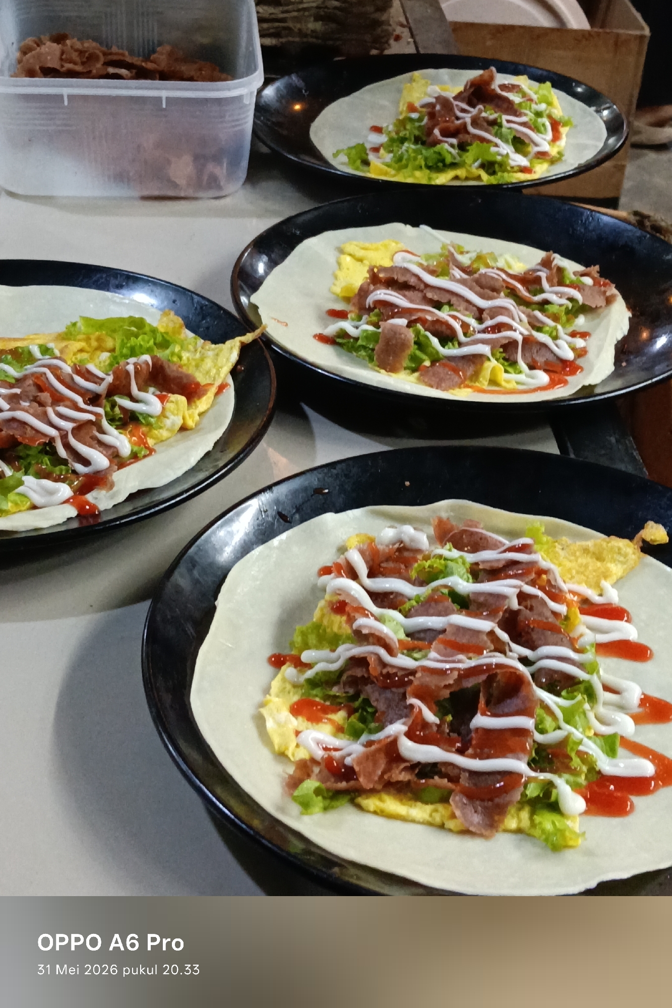 The image depicts a culinary scene featuring four plates of what appear to be open-faced shawarma wraps. Each wrap consists of a thin, pale tortilla base topped with scrambled eggs, shredded lettuce, slices of meat (likely shawarma), and drizzled with both ketchup and a white sauce, possibly mayonnaise or a similar condiment. The plates are black and have a glossy finish. In the background, a clear plastic container filled with sliced meat is visible. The environment appears to be an outdoor dining area, possibly in Sangso, Indonesia, as requested, though this cannot be definitively confirmed from the image alone. No people are visible in the frame.