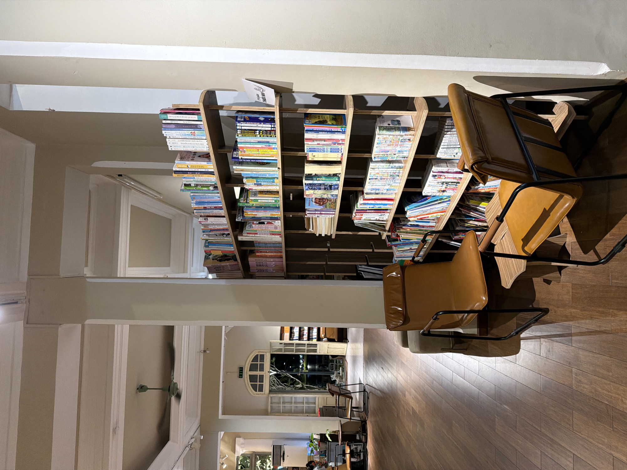 A dimly lit interior of what appears to be a cafe or bookstore in Thuận Hóa Ward, Vietnam, features a tall, multi-shelf wooden bookcase filled with various books. Two brown leather armchairs with dark metal frames are positioned in the foreground, one closer to the camera and slightly to the right, the other further back and to the left. The floor is made of dark, polished wooden planks. Through an open doorway in the background, a nighttime outdoor scene is visible, with lights and foliage. A ceiling fan is visible in the upper left portion of the image.