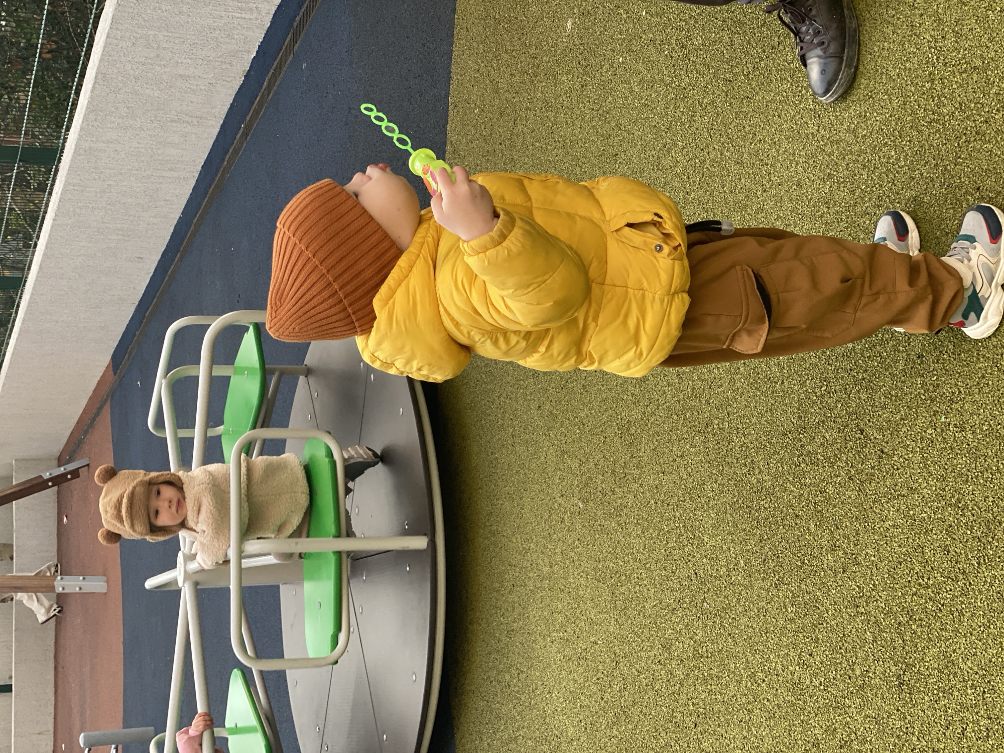 The image shows two children at a playground. One child, wearing a brown beanie, yellow puffer jacket, and brown pants, stands on a green rubber ground surface and is blowing bubbles. The second child is sitting on a small, merry-go-round, wearing a light brown bear-eared hooded coat. A person's black shoe is visible in the top right corner. The playground has areas of blue and gray surfaces. The surrounding area includes trees and building structures. The specific location in Sochi, Russia cannot be definitively determined from the image alone.