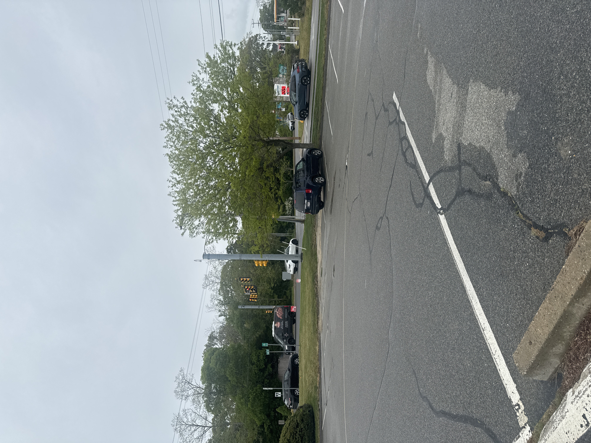 The image shows a street scene in Eastham, United States of America. The photo appears to have been taken from a parking lot.
Visible features:
- An asphalt parking lot with painted white lines and cracks.
- A curb.
- Several parked cars, including dark-colored SUVs and a white sedan.
- A green tree.
- A gas station sign displaying a price of "$4.69".
- A traffic light with yellow lights.
- A tree line, utility poles and wires.
- A gray, overcast sky.