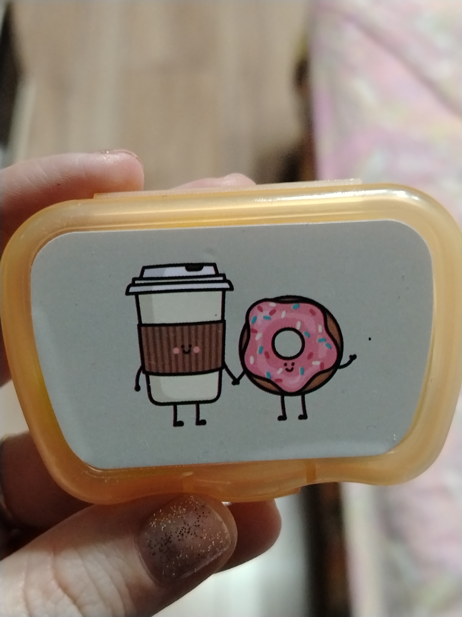 A close-up shows a small, rectangular, light-orange container held in a person's hand. The container has a light-gray top with an illustration of a cartoon coffee cup and a donut holding hands. The coffee cup is white with a brown textured band, a black lid, a small smiling face, arms and legs. The donut is pink with sprinkles, a brown ring, a smiling face, arms, and legs. The person's fingernail visible is painted with glittery nail polish. The background is blurred, showing a light-colored wooden surface and a pink and white patterned object.