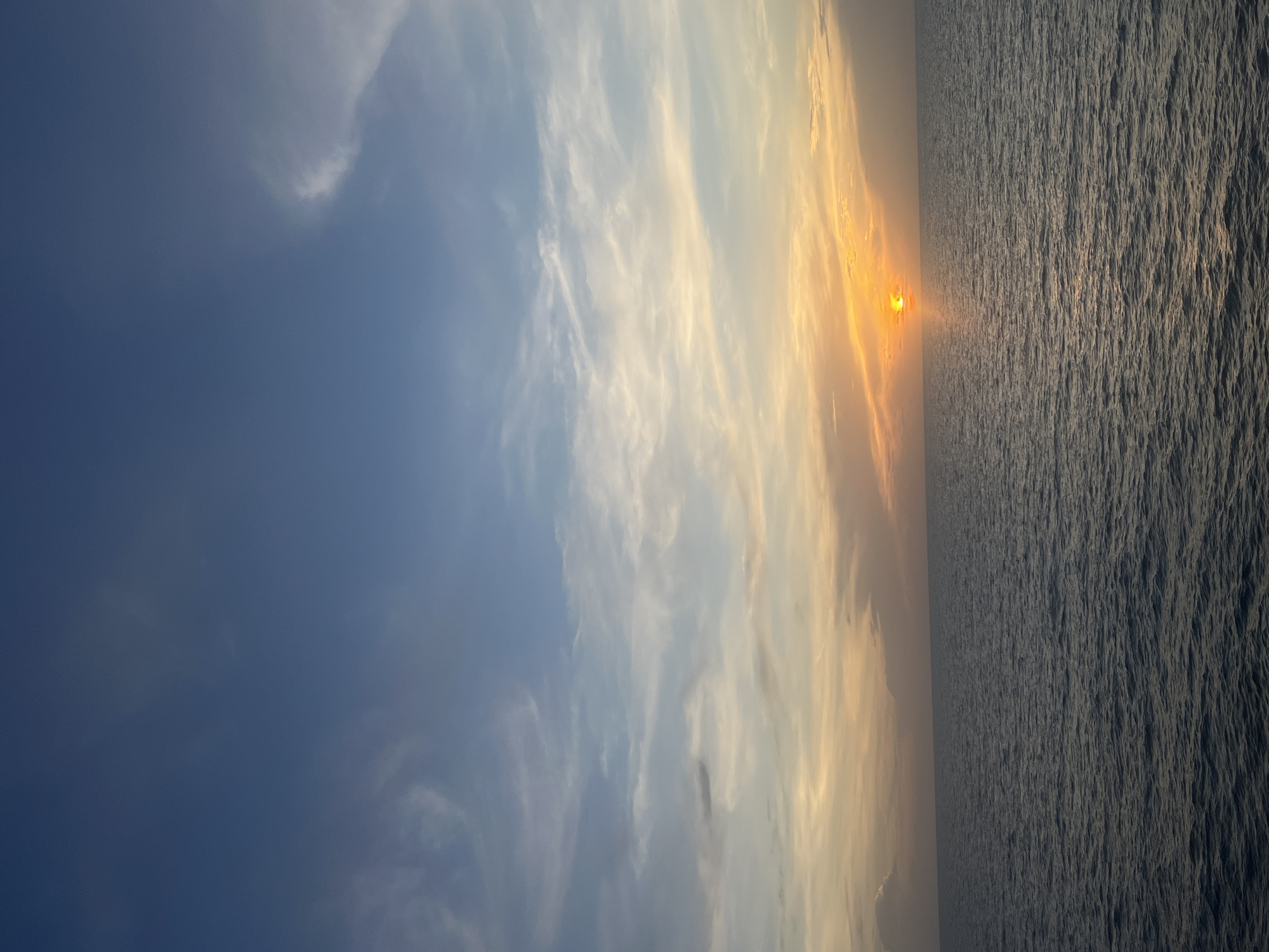 The image captures a horizon view, with the sky transitioning from a deep blue on the left to lighter hues and wispy clouds towards the center and right where the sun is setting. A sliver of bright orange sun is visible on the horizon, reflecting on the ocean water. The sea occupies the right portion of the image, its surface reflecting light. The location is Unknown, Unknown.