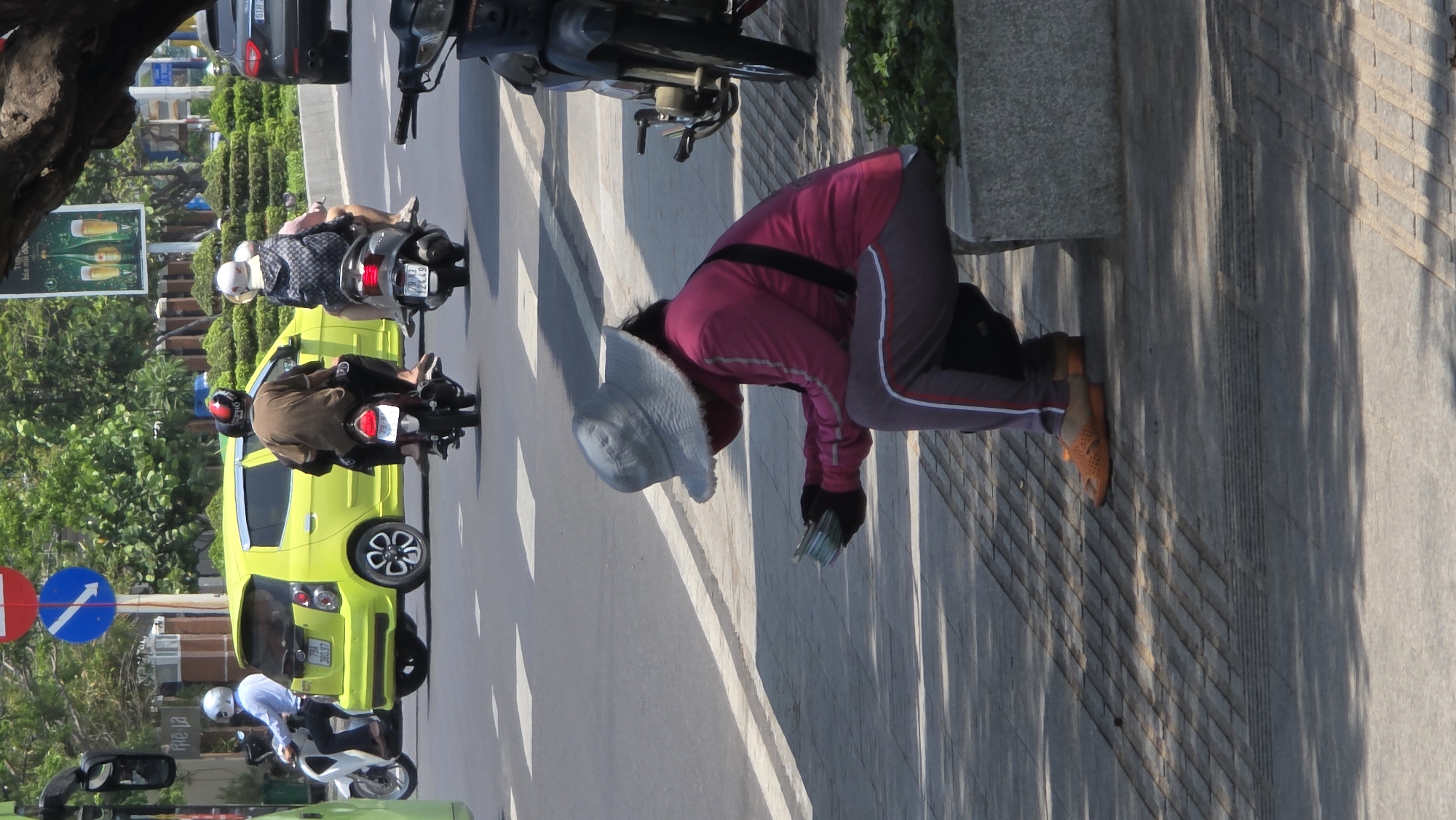 An eye-level, medium shot of a street scene in Nha Trang, Vietnam, showing pedestrians, motorbikes, and vehicles, alongside a roadside stone wall. The photograph is oriented with the street to the left side of the image and the pedestrian side to the right side.
In the right of the frame, a woman wearing a light-colored sun hat, a pink jacket with dark sleeves, and grey pants with a stripe down the side is bent over, appearing to pick something up from the walkway next to a low stone wall. She's wearing orange sandals or shoes.
To the left of her is a street with a variety of vehicles. Near the top of the street, there's a black car, and in front of that, a parked motorbike. Further down the street, there are several motorbikes with riders wearing helmets, and behind them, a small lime-green hatchback car. A sign with blue and red circles indicates traffic regulations.
The walkway is paved with light-colored concrete or stone, and long shadows stretch across it from left to right. A tree branch is visible in the upper left corner of the frame. A roadside advertisement is seen on the left.