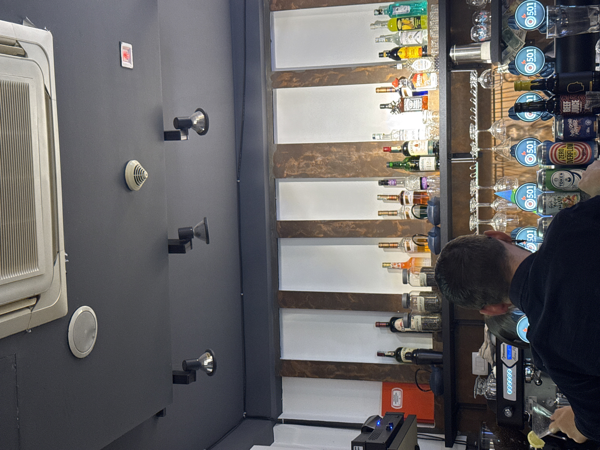 Here is a detailed description of the image:
**Setting:** This image captures the interior of a bar, as indicated by the bar shelves filled with bottles of alcohol and cocktail ingredients. The location in the city of Bar, Montenegro, is indirectly confirmed by the repeated presence of the logo "501 BARTS BAR," which is presumably the name of the establishment.
**Objects:** The main visual elements include:
* Shelves laden with a variety of alcoholic beverages. Specific brands visible include Jameson and Kahlua. Cocktail glasses also line the shelves.
* A bartender, engaged in preparing a drink.
* Bar equipment. This includes what seems to be an espresso machine, and other tools required for preparing cocktails.
* Architectural details like a wall with spotlights, a fire alarm, and an air conditioning unit, all painted in gray. There is also a switch on the wall.
**People:**
The bartender is partially visible, likely a male with short dark hair wearing a black shirt. His focus is on the preparation of a drink.
**Action:**
The primary action is the bartender preparing a drink.
**Environment:** The interior is likely a modern or contemporary bar. The gray walls give a sleek appearance. The bottles and equipment suggest a place where a variety of drinks are served.
In essence, the image depicts a bartender in a bar setting, potentially at the "501 BARTS BAR" located in Bar, Montenegro, occupied with the task of mixing a drink.