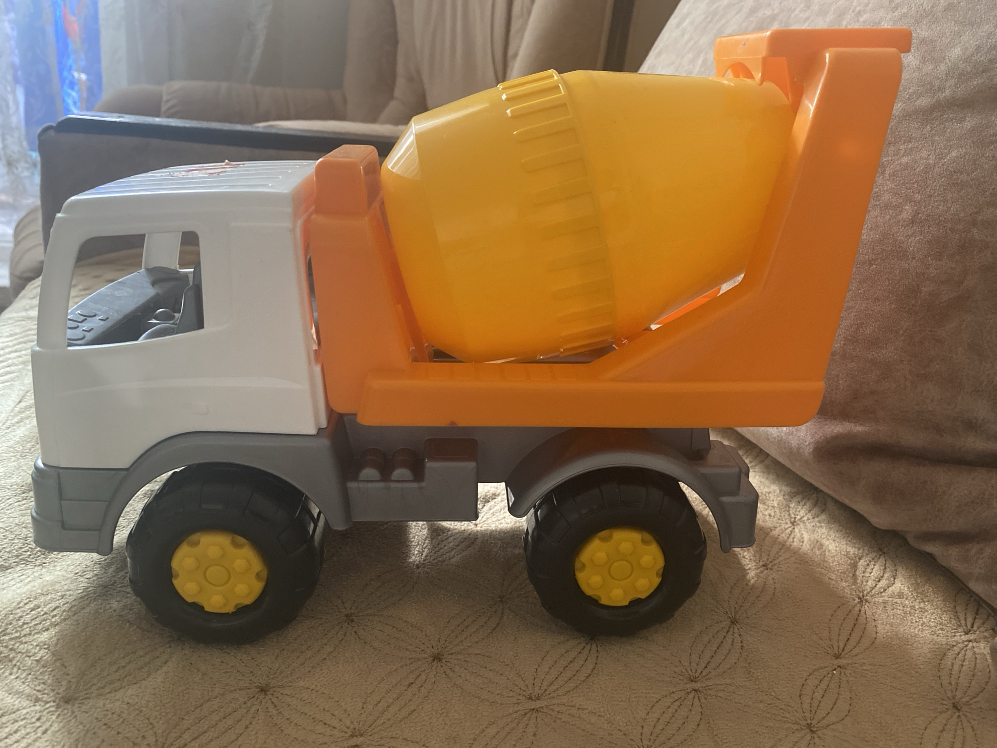 A toy cement mixer truck is positioned on a textured, light brown fabric surface, likely a couch or bed. The truck is primarily white and orange, with a yellow drum. The truck has four black wheels with yellow hubs. The background shows a blurred indoor environment with furniture. The image does not provide any information about the city of Kizilyurt, Russia, or any specific location within it.