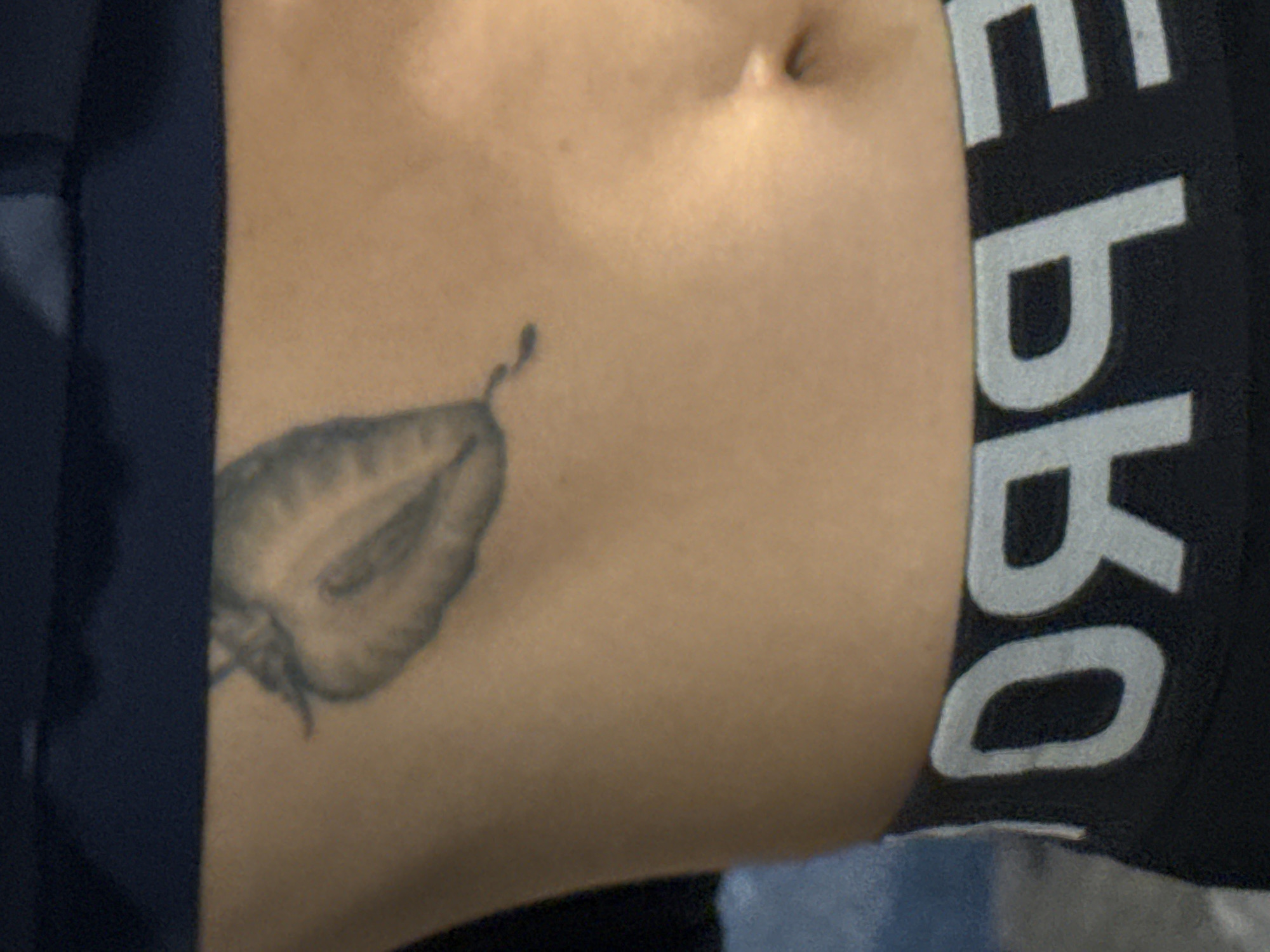 A close-up shot shows a person's midsection. A strawberry tattoo is on the lower left abdomen. The navel is visible near the top center of the frame. A dark-colored garment with white lettering is partially visible on the right side of the image.