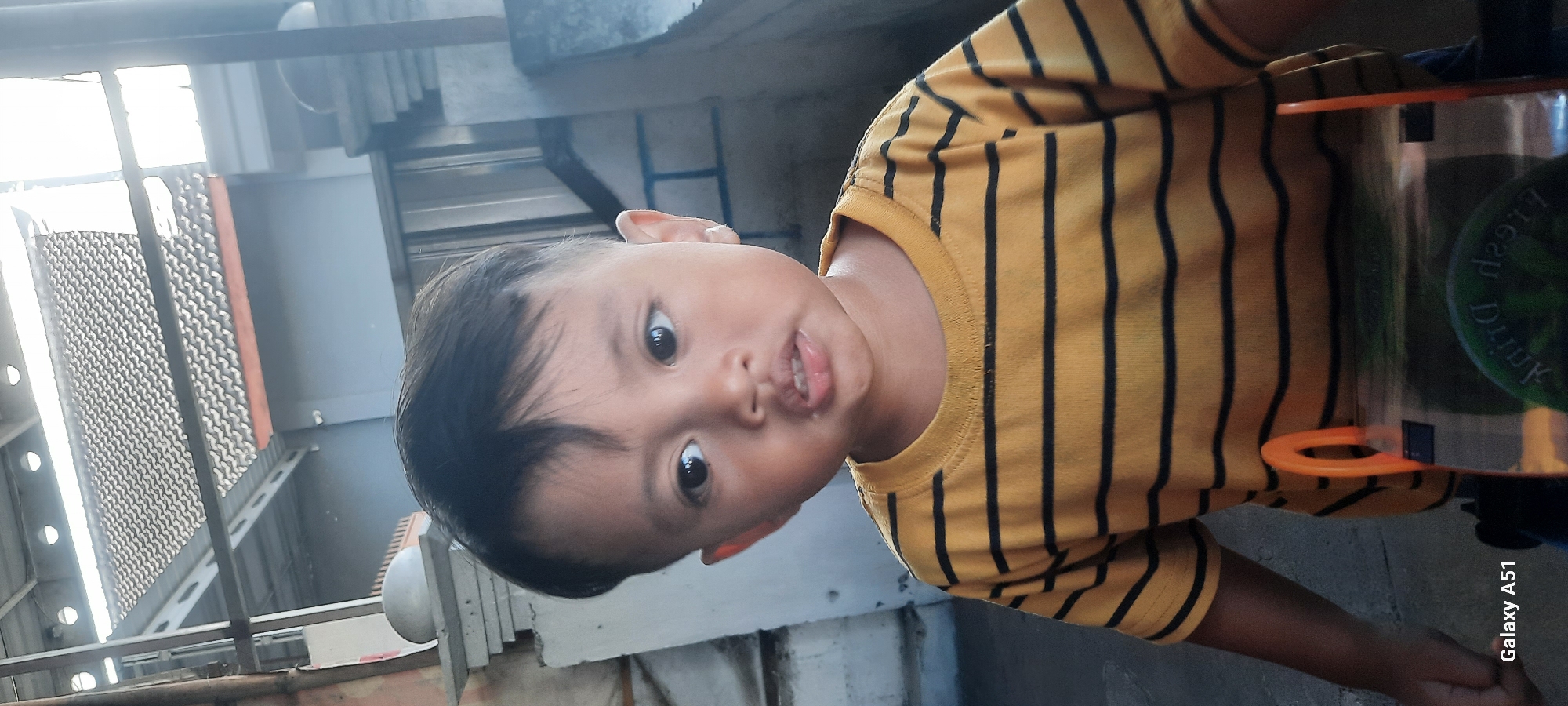 A young boy with dark hair and large, dark eyes is facing forward, with his head slightly tilted. He has light-colored skin and appears to be wearing a yellow t-shirt with black horizontal stripes. His mouth is slightly open, revealing his teeth and tongue. In the background, there is a building with a textured roof and some vertical supports. The lighting suggests it is daytime. The bottom left corner of the image displays the text "Galaxy A51". The scene is likely an outdoor setting in Selopuro, Indonesia.