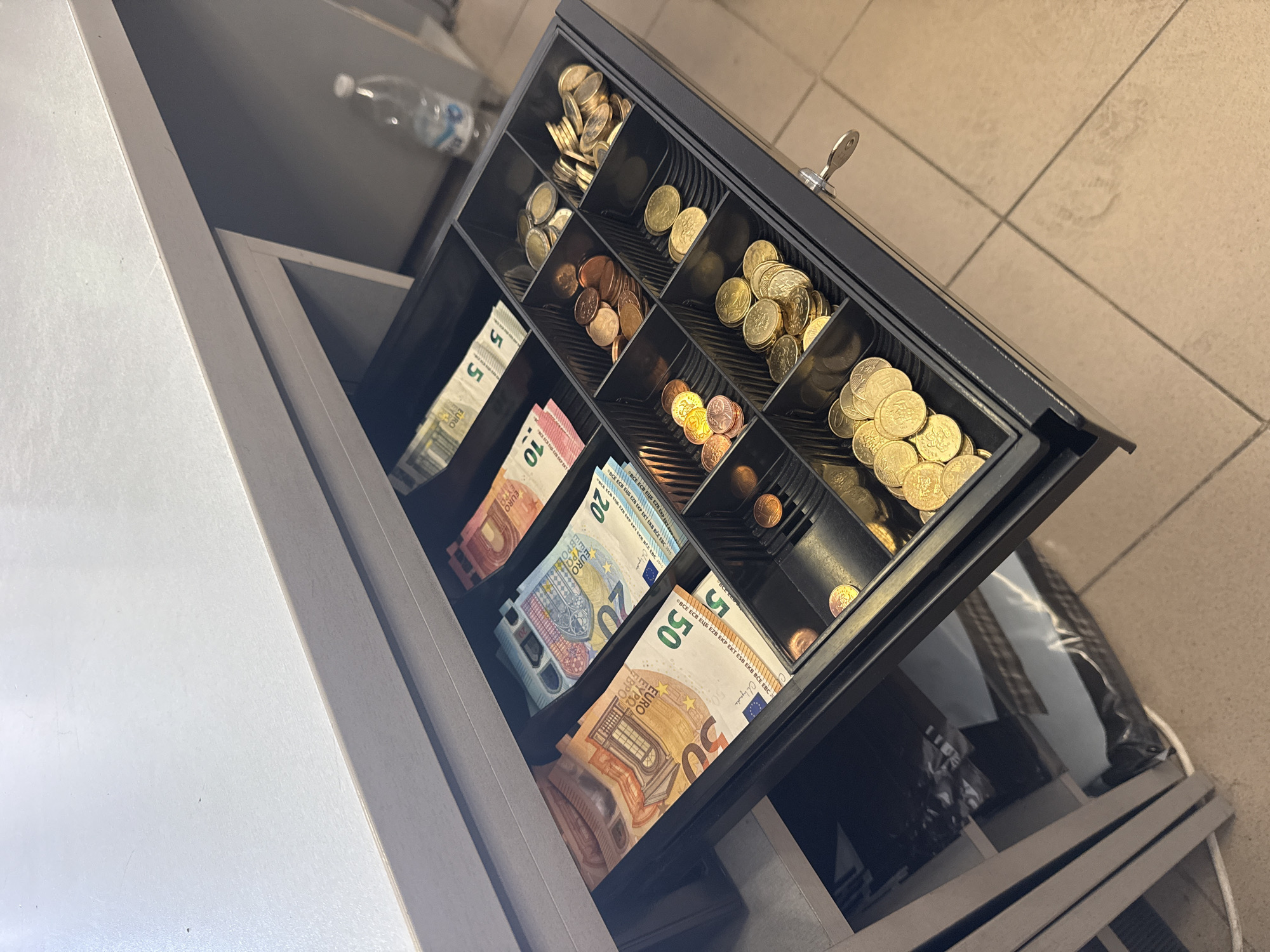 A cash drawer is open, revealing euro banknotes and coins. The banknotes are denominations of 50, 20, and 5 euros, neatly stacked in their respective compartments. The coins are various denominations and are in separate compartments as well, some appearing to be copper-colored and others gold-colored. The drawer is made of black metal and has a key inserted into its lock on the right side. The drawer is part of a counter, which appears to be made of a light-colored material. In the background, a partially visible plastic bottle of water and some shelves are present. The floor is tiled with a pattern of light and dark beige squares. The scene is indoors and appears to be in a commercial establishment, possibly a store or restaurant. The image was taken in the city of Rakovski, Bulgaria.