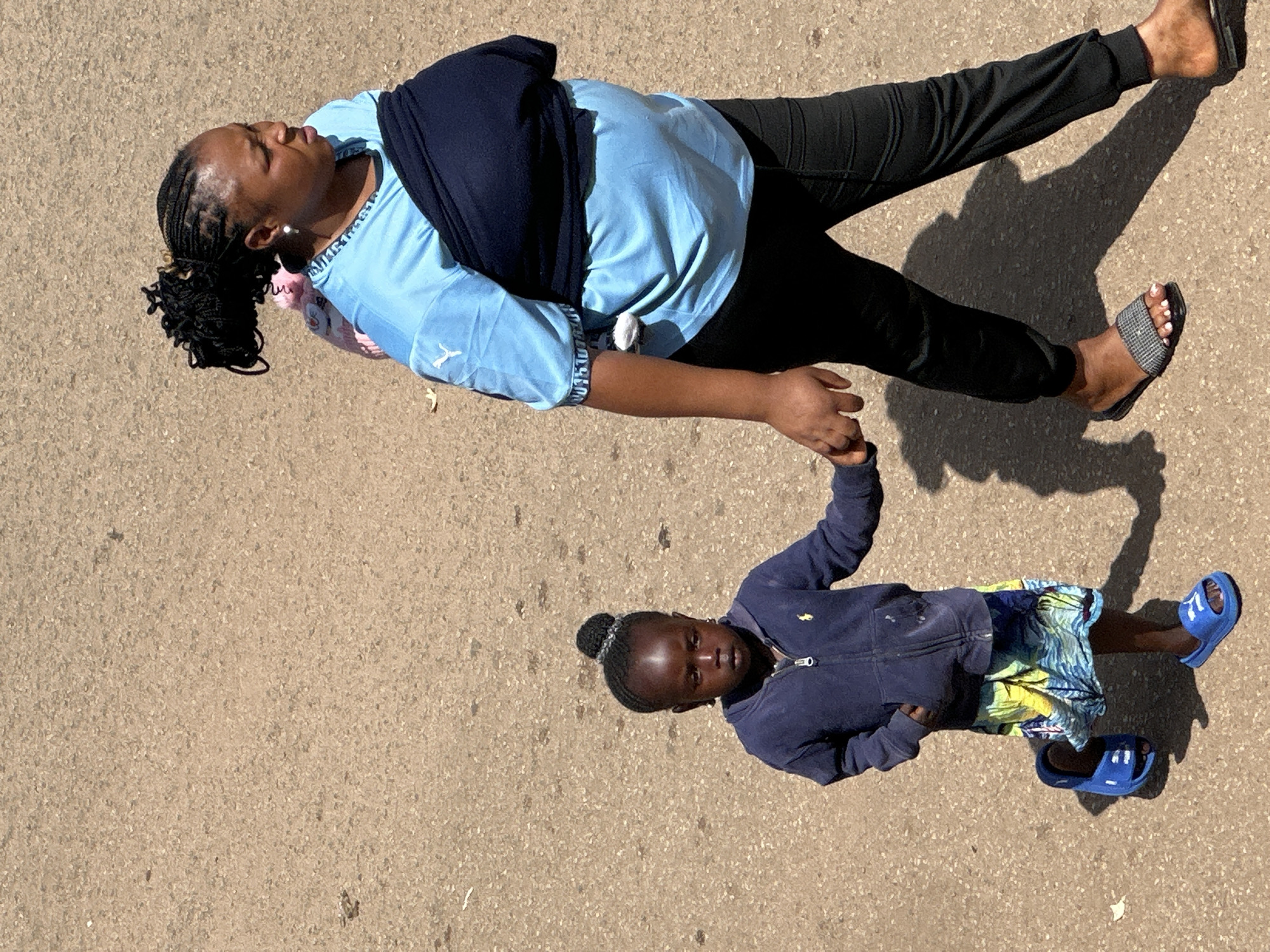 In the city of Bukuru, Nigeria, a woman carrying a baby on her back walks alongside a young child. The woman is wearing a light blue t-shirt and black pants, with a dark blue wrap around her waist and the baby. The child is wearing a dark blue hooded jacket and a colorful skirt, with blue sandals. They are walking on a paved surface, likely a road or sidewalk, with shadows cast by the sun. The overall scene depicts a common urban environment with everyday activity.