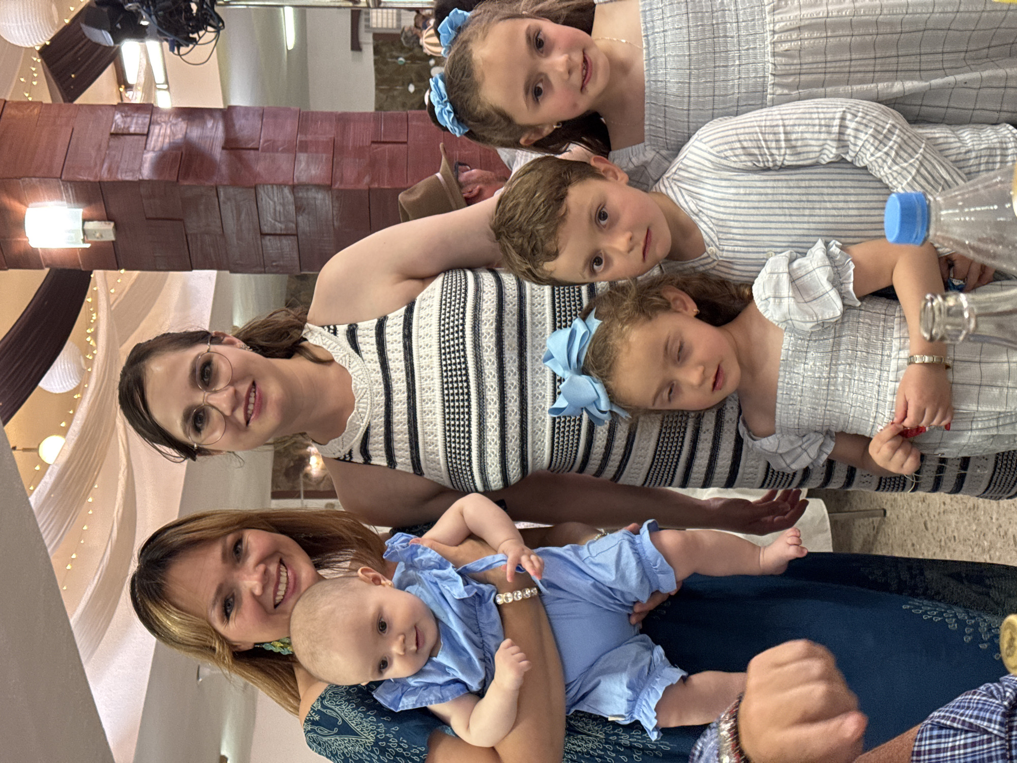 A group of seven people pose for a photograph indoors. From the bottom up, a woman with blonde hair smiles and holds a baby dressed in blue. Behind them, a woman with glasses smiles, and behind her are three children wearing blue and white striped clothing and blue bows. A water bottle is also visible. A man wearing a hat is partially visible behind the woman with glasses. The location appears to be a celebratory event, suggested by the decorations in the background, possibly in the city of Chipilo de Francisco Javier Mina (Chipilo), Mexico.
