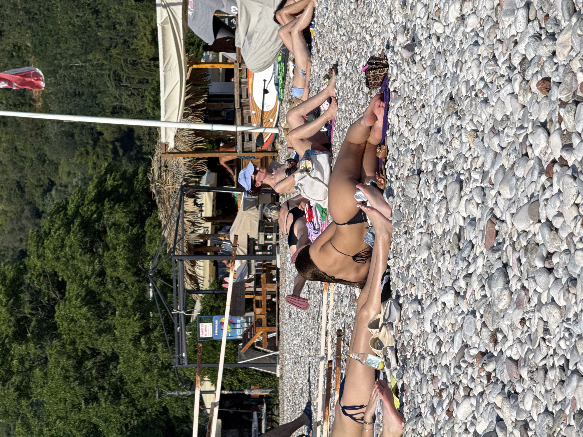 This media file captures a sunny day at a pebble beach in Budva, Montenegro. The scene is populated with several individuals relaxing on the shore, some sunbathing, others sitting or lying down. The foreground is dominated by the textured surface of numerous grey and white pebbles.
Several people are dressed in swimwear, suggesting a leisure activity at the beach. One person in the foreground is lying on their side, exposing their back, while another is seated. Further back, more figures are visible, some partially obscured by others or by beach structures.
Notable details include a white and red flag or banner on a pole in the upper left of the frame. Beach umbrellas and thatched-roof structures are visible in the background, indicating a developed beach area with amenities. A stand-up paddleboard is propped up against some beach furniture. The presence of sunglasses, towels, and a bottle of water further accentuates the beach setting. The lighting suggests a bright, daytime scene, likely late morning or afternoon, with clear skies and warm weather.
A sign with text is partially visible in the middle ground. One part of the sign has the text "TRANSFER" and "V NIKOLA". Another sign below that appears to have "PARADISE" written on it. These signs might indicate local services or a place name. The overall mood is relaxed and leisurely, consistent with a holiday or vacation at a seaside resort.