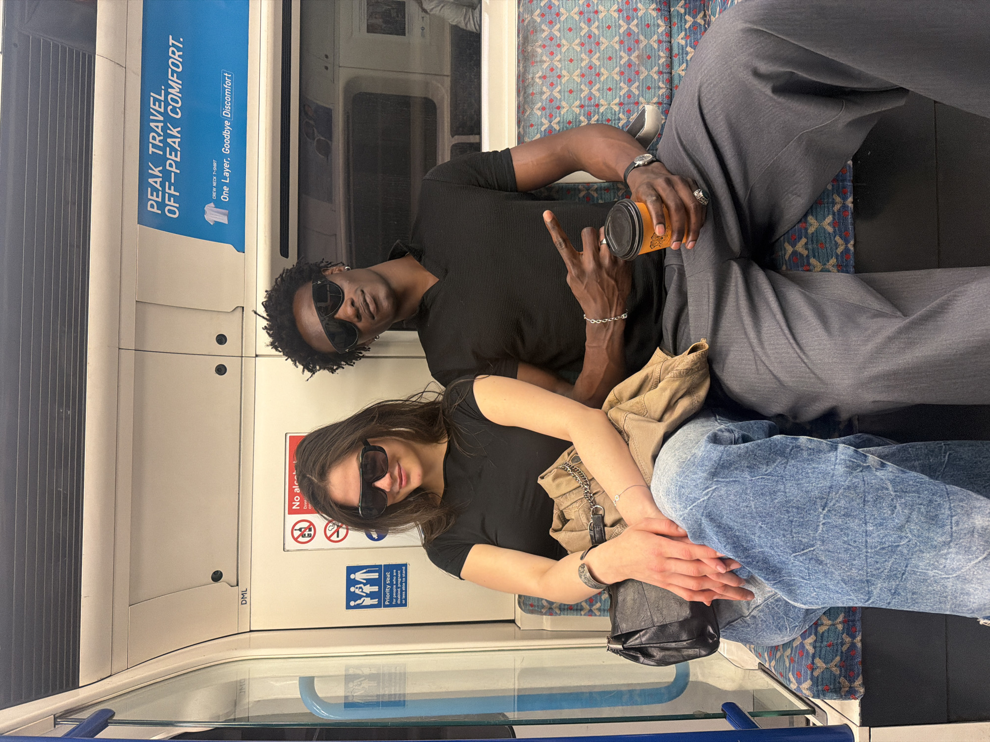 Two individuals, a man and a woman, are seated side-by-side in what appears to be a London Underground train carriage. The man, wearing sunglasses and a black t-shirt, is holding a beverage cup and gesturing with his hand. He also wears a wristwatch and jewelry. He is sitting to the left. The woman, also in sunglasses and a black t-shirt, has her hands resting on her bag. She is sitting to the right. Both individuals are wearing sunglasses. The train carriage interior features signs and advertisements, including a "Priority seat" sign and an advertisement promoting "PEAK TRAVEL. OFF-PEAK COMFORT." The seat upholstery has a distinctive pattern. The location is London, United Kingdom, specifically inside a London Underground train.