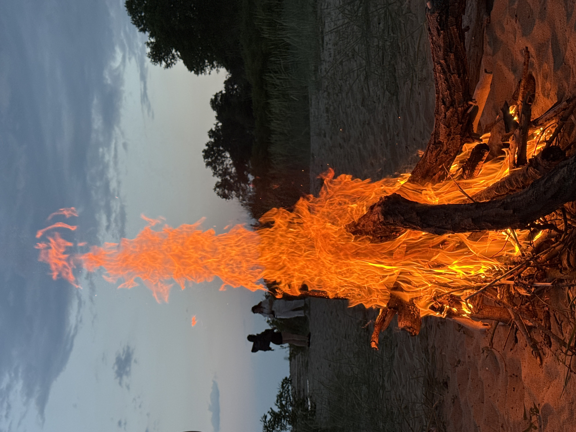 A bonfire burns on a sandy beach at dusk. The flames are high and bright orange, consuming several branches. Two people stand in the background, slightly obscured by the flames. The sky is a pale blue-gray, and there is vegetation along the shoreline in the background. The location is likely near Novo Oriahovo, Bulgaria, given the prompt's information.