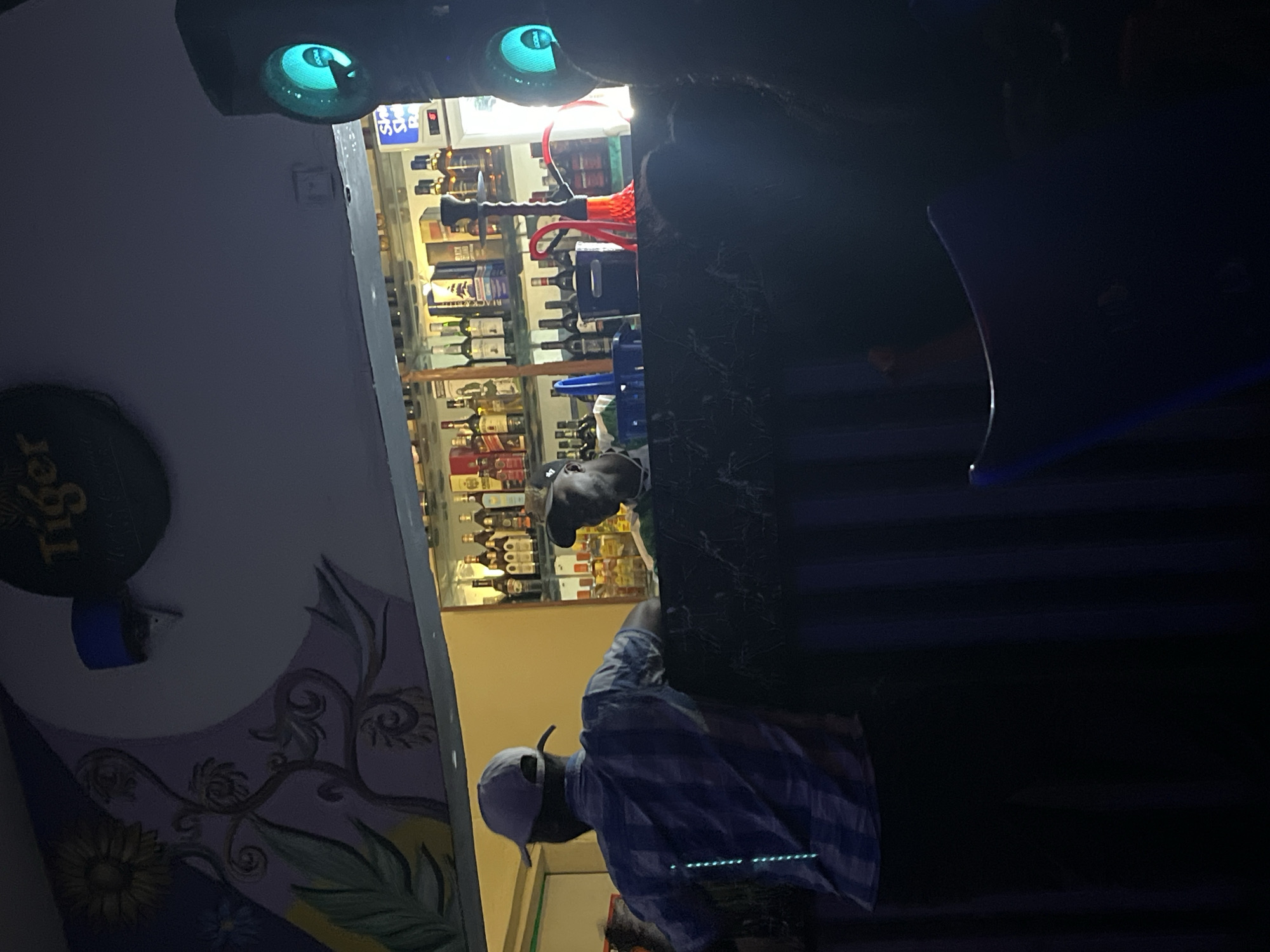 This image depicts the interior of a dimly lit bar or lounge in Jos, Nigeria. A person wearing a blue and white checkered shirt and a light-colored baseball cap is standing with their back to the camera, facing a bar. Behind the bar, shelves are stocked with various bottles of liquor. Another person, wearing a dark baseball cap, is visible behind the bar. A red hookah pipe is prominently displayed on the bar. The overall ambiance is that of a social establishment with a focus on drinks and potentially shisha. The lighting is moody, with neon or LED lights visible in some areas.
