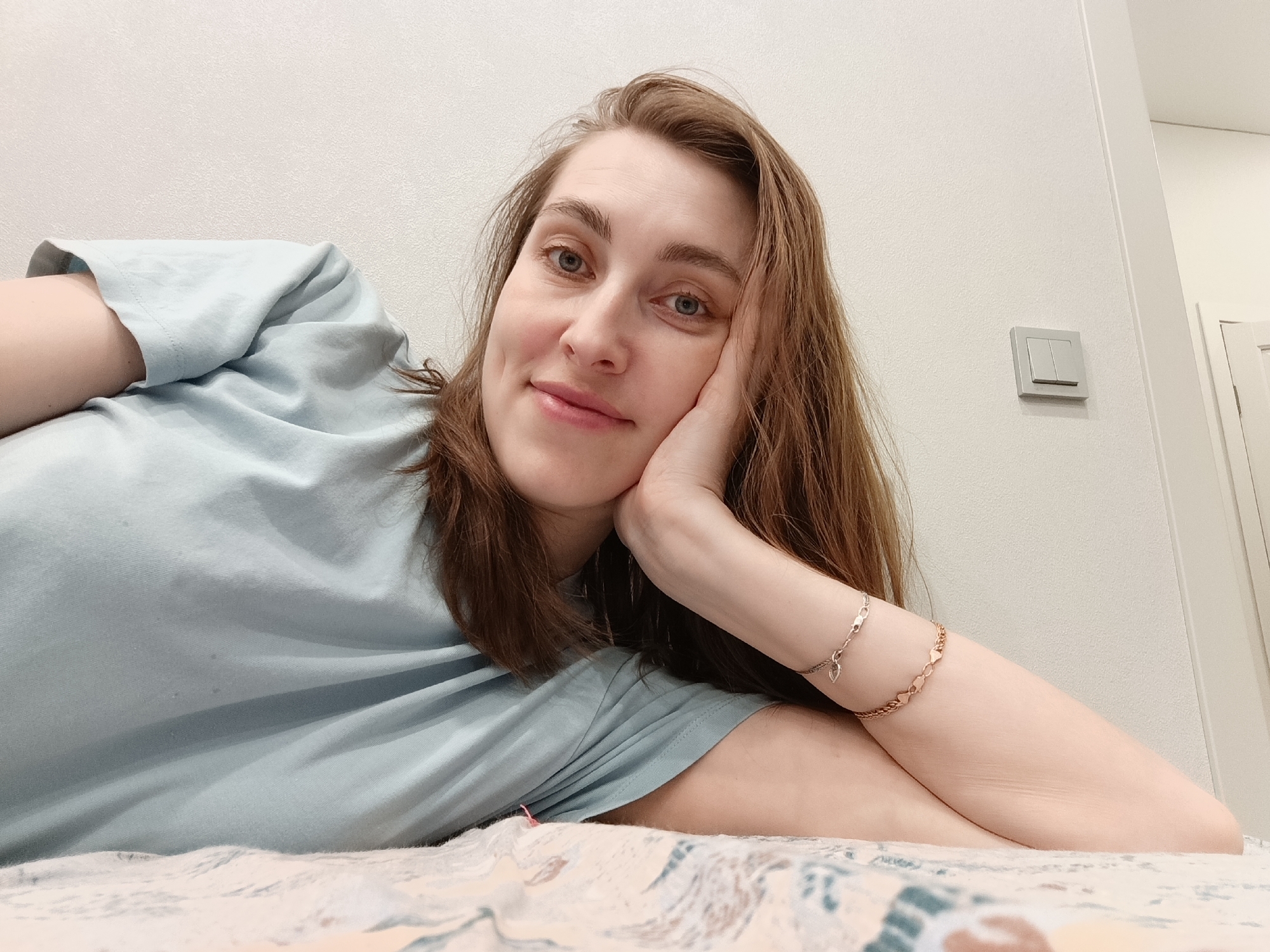 A woman is lying on a bed in what appears to be an indoor setting. The woman has light skin, blue eyes, and light brown hair. She is wearing a light blue t-shirt and two bracelets on her right wrist. She is propped up on her left side with her right hand resting against her face. The background consists of a white wall with a light switch and the edge of a doorway visible. The bed has a patterned bedspread. The location is Chaikovsky, Russia.