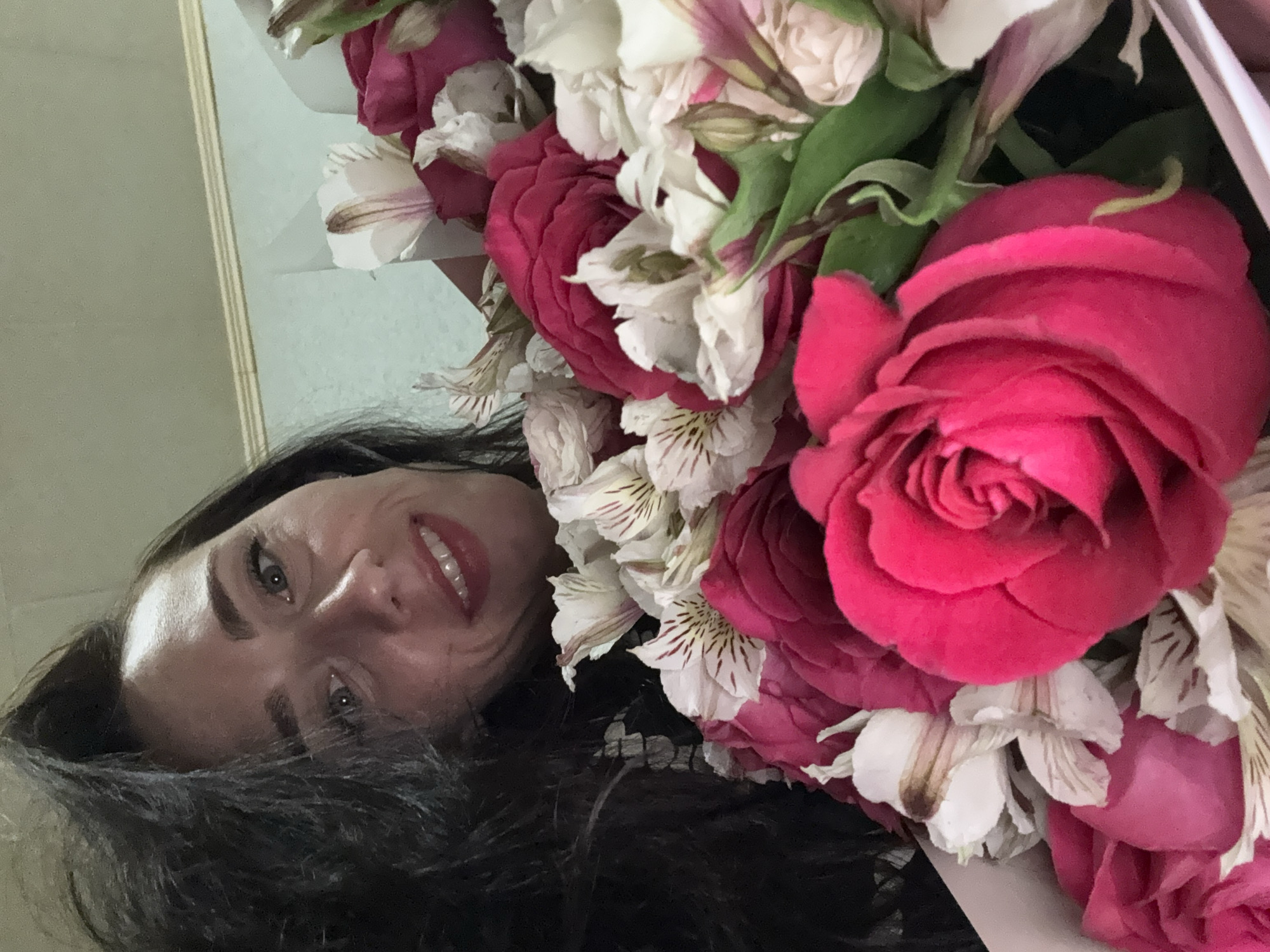 A person with dark hair is holding a bouquet of red and white flowers, partially obscuring their face. The person's face is visible, with fair skin, grey eyes, and a smile. The background appears to be a light-colored wall, potentially indoors. There is no indication of the location being Saint Petersburg, Russia.