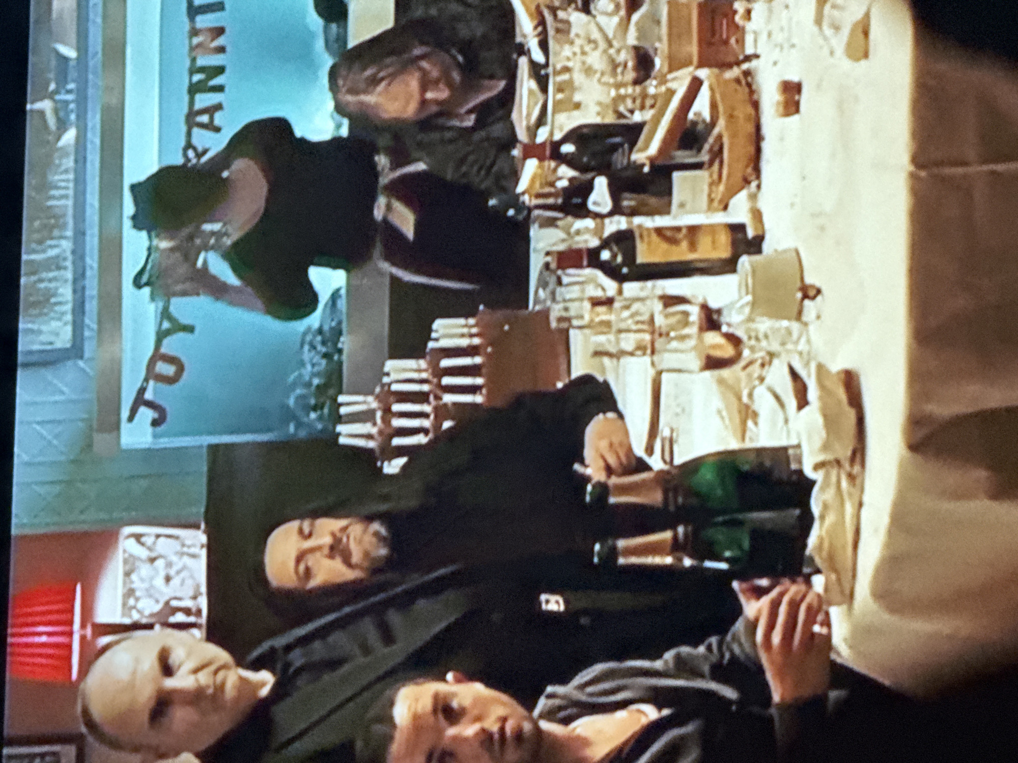 The image depicts a gathering, possibly a celebration, with several people and a table laden with food and beverages. There are at least three men visible in the foreground, one with a shaved head. On the table, there are bottles of wine and champagne, as well as numerous glasses. In the background, a woman is seen holding up a glass, possibly in a toast. A banner with the word "ANNIVERSARY" is displayed. There is no information on the location in the city of Mytishchi, Russia.
