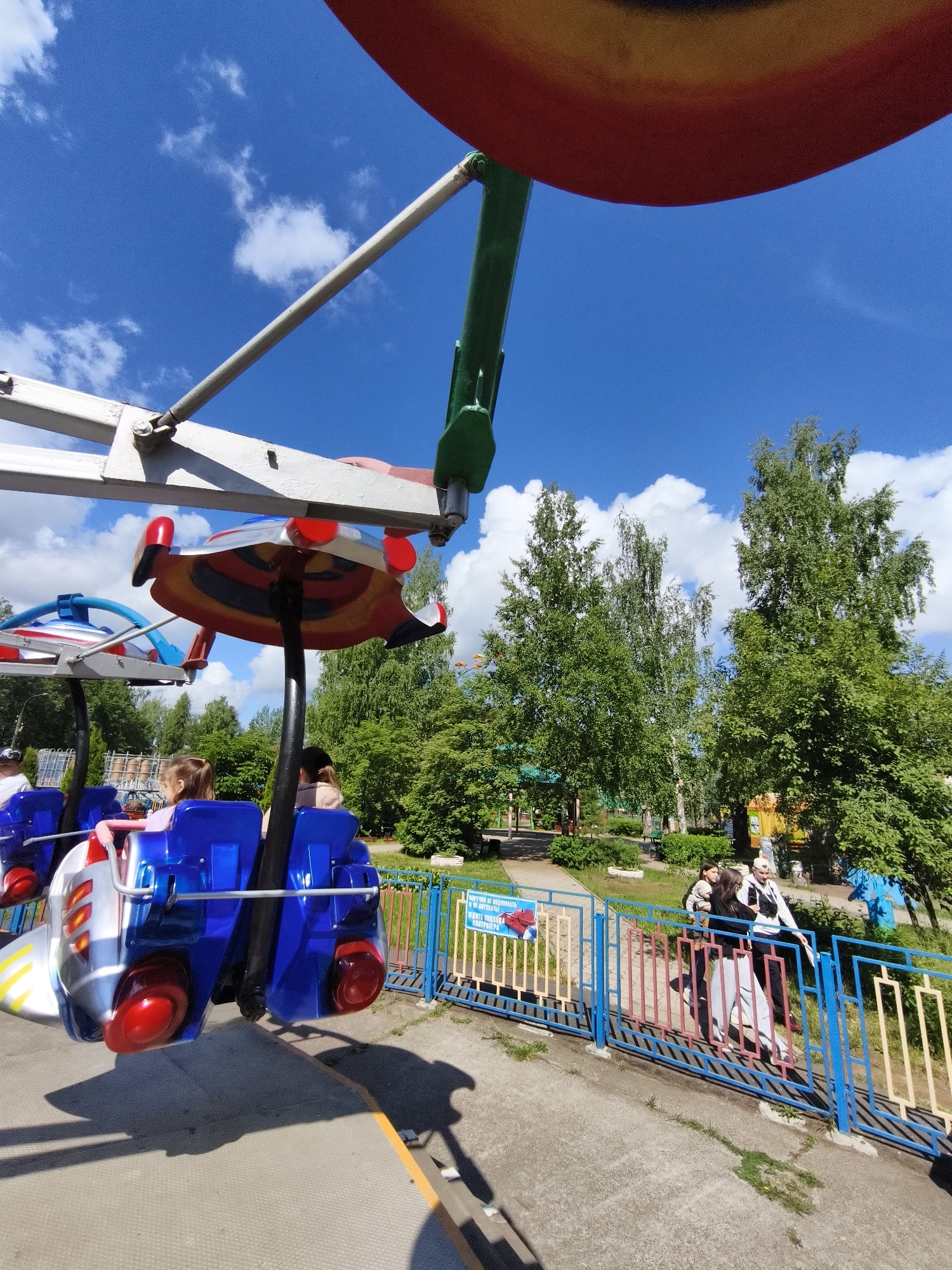 The image shows a children's amusement ride in Chaikovsky, Russia. The ride has several blue "airplane" seats suspended from a colorful rotating structure. Children are seated in some of the airplanes. A metal railing separates the ride from a walkway where a small group of people is standing. Trees and greenery are visible in the background under a blue sky with white clouds.
