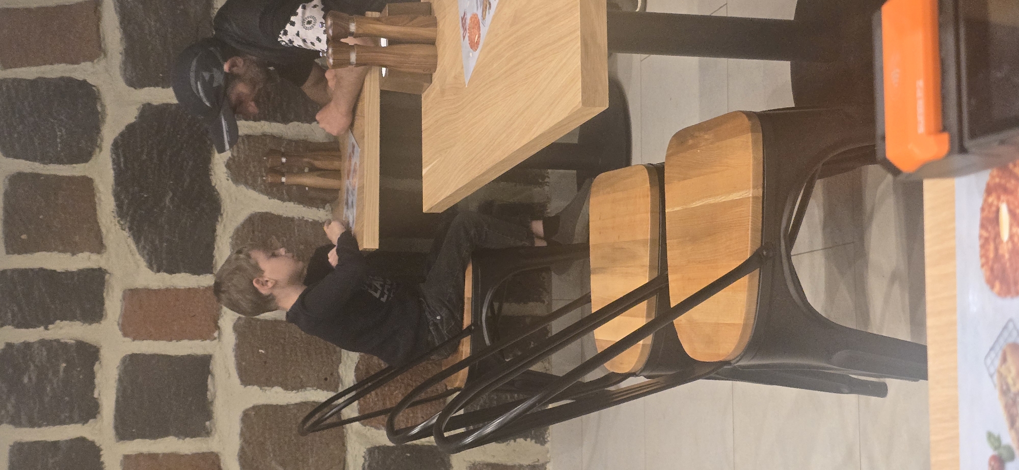 An indoor scene shows a man and a young boy sitting at a light-colored wooden table. The man, seen from the shoulders up, wears a black cap and a black and white patterned shirt. The boy, sitting at the table with his back to the camera, wears a black shirt and pants. The table has what appears to be menus or papers on it. There are wooden salt and pepper shakers on the table. The scene is set indoors, potentially in a restaurant in Yerevan, Armenia, based on the visible style of the stone floor. The seating consists of light-colored wooden chairs with black metal frames. An orange dispenser is visible on the right side of the image near a promotional sign.