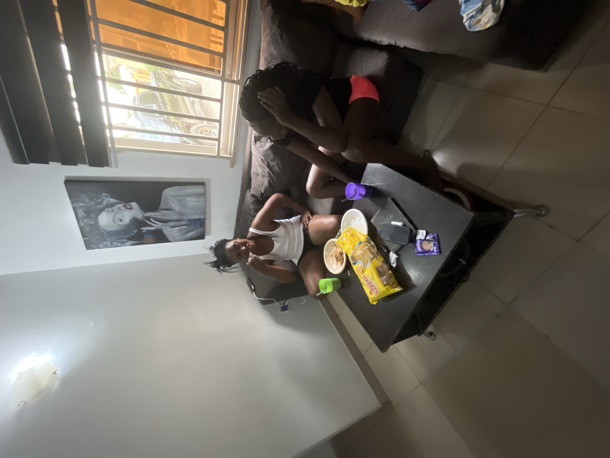 In this indoor scene, two young Black women are relaxing on a couch in what appears to be a living room. The woman in the foreground is seated and wearing a white tank top and black shorts, eating from a bowl. She is looking towards the camera with a neutral expression. Next to her on the floor is a green cup. Beside her on the coffee table is a yellow bag of snacks and a purple cup.
The second woman is seated on the couch behind the first, leaning back and covering her face with her hands, her pose suggesting distress or discomfort. She is wearing a black t-shirt and pink shorts.
On the coffee table, there are several items including a wrapped candy, a phone, and the aforementioned snacks and cups. Above the couch, on the wall, hangs a framed black and white print of a woman's face.
To the left of the frame, a window with blinds and bars is visible, allowing bright daylight to stream in, suggesting it is daytime. Outside the window, a car is visible. The room is furnished with a dark sofa and a dark coffee table. The floor is tiled. The overall mood seems relaxed, though the second woman's posture introduces a note of concern. The location is identified as Abuja, Nigeria.