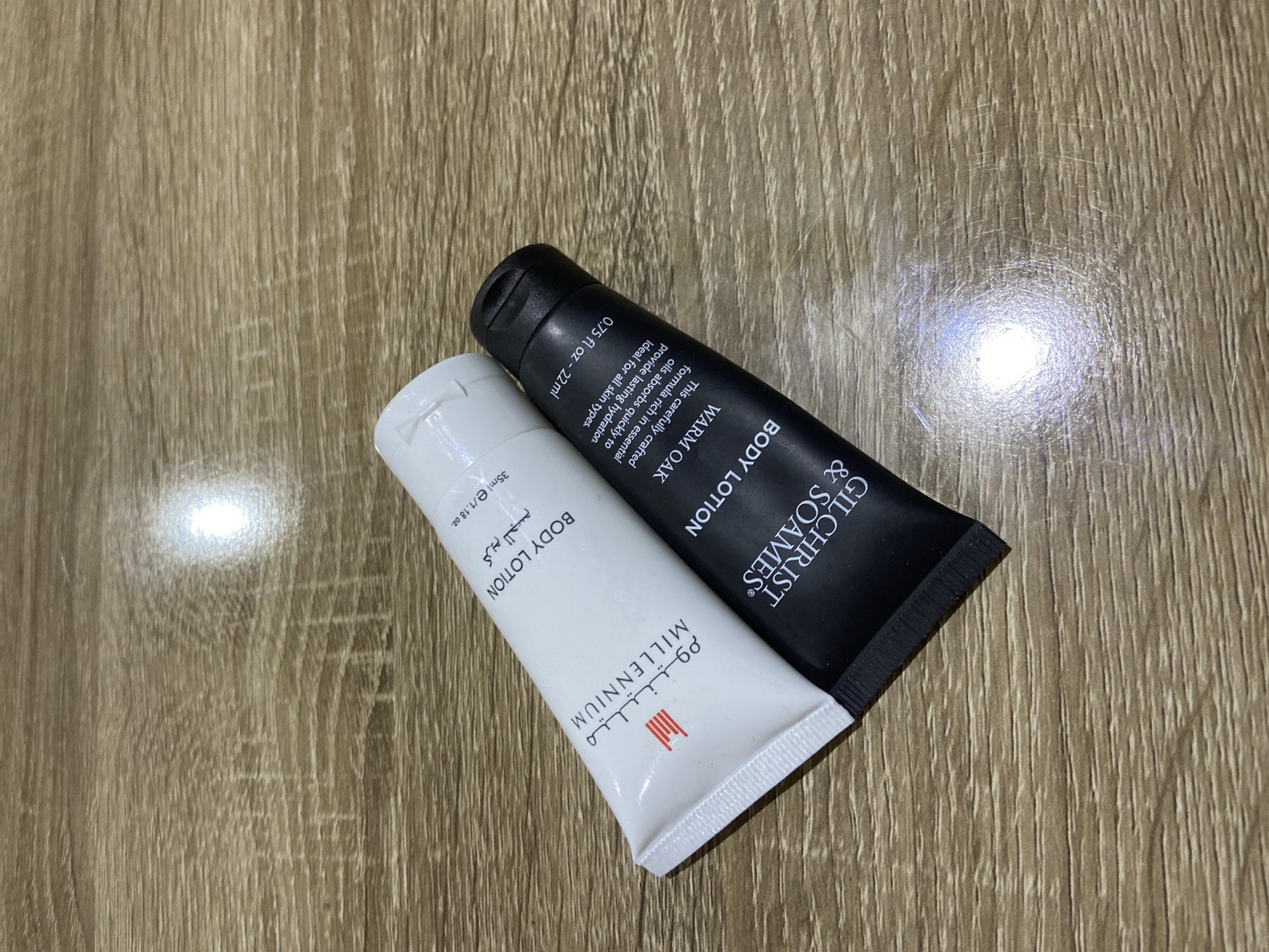 Two travel-sized tubes of body lotion lie on a light brown wooden surface. The tube on the left is white with black text and Arabic script. The text on the white tube reads "MILLENNIUM BODY LOTION" and "35ml e/1.18 oz". The tube on the right is black with white text. The text on the black tube reads "GILCHRIST & SOAMES BODY LOTION WARM OAK" and "0.75 Fl oz - 22 ml". The tubes are positioned diagonally, with the white tube slightly overlapping the black tube. The lighting creates a bright reflection on the wooden surface. The location is not specified in the image or provided context.
