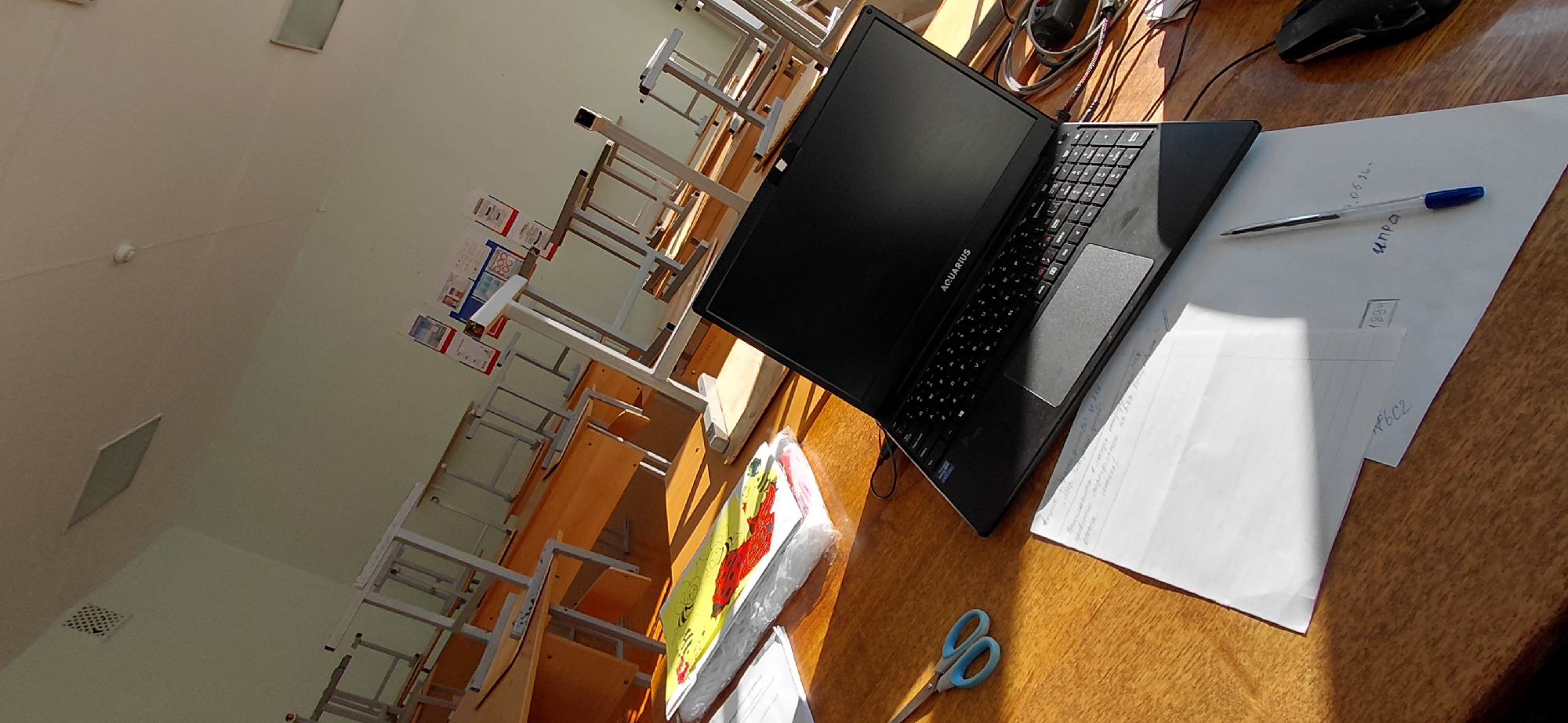 This is a medium shot, taken from a slightly elevated angle, of a classroom desk in Svetlogorsk, Russia. The desk is made of wood and is positioned in front of a row of identical student desks with metal frames and wooden surfaces. The classroom appears to be empty, and the desks are stacked in a way that suggests they are not in use. The lighting suggests it is daytime, with sunlight streaming in from the right side of the frame, casting long shadows across the desk.
On the desk in the foreground, there is an open black laptop with the brand name "AQUARIUS" visible on the lid. To the left of the laptop, a pair of blue scissors rests on a stack of papers and a colorful children's book. To the right of the laptop, a computer mouse is partially visible, connected by a black cable. In front of the laptop, a white sheet of paper is placed, with a blue ballpoint pen lying on it. Some handwritten notes are visible on this paper, as well as on another sheet of paper underneath. In the background, posters are displayed on the wall, and there are ventilation grilles and a smoke detector on the ceiling. The overall scene conveys a sense of an academic environment, possibly a classroom after school hours or during a break.