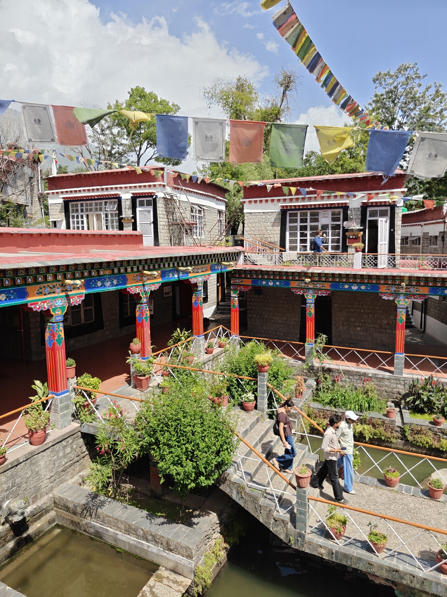 The image captures a courtyard scene at the Norbulingka Institute, a Tibetan cultural center in McLeod Ganj, near Fatehpur, India. Two low-rise stone buildings with red roofs and Tibetan architectural details surround the courtyard. Rows of colorful prayer flags are strung overhead.
The courtyard features a water feature crossed by a stone bridge with white railings and potted plants. Three people are walking across the bridge: a woman in jeans and a patterned shirt, a man in black pants and a light-colored shirt, and a man in blue pants and a white cap. The surrounding area is landscaped with various plants and flowers. The columns supporting the buildings' overhanging roofs are painted in vibrant colors and patterns. A man is seen on the balcony of one of the buildings. The sky is partly cloudy.