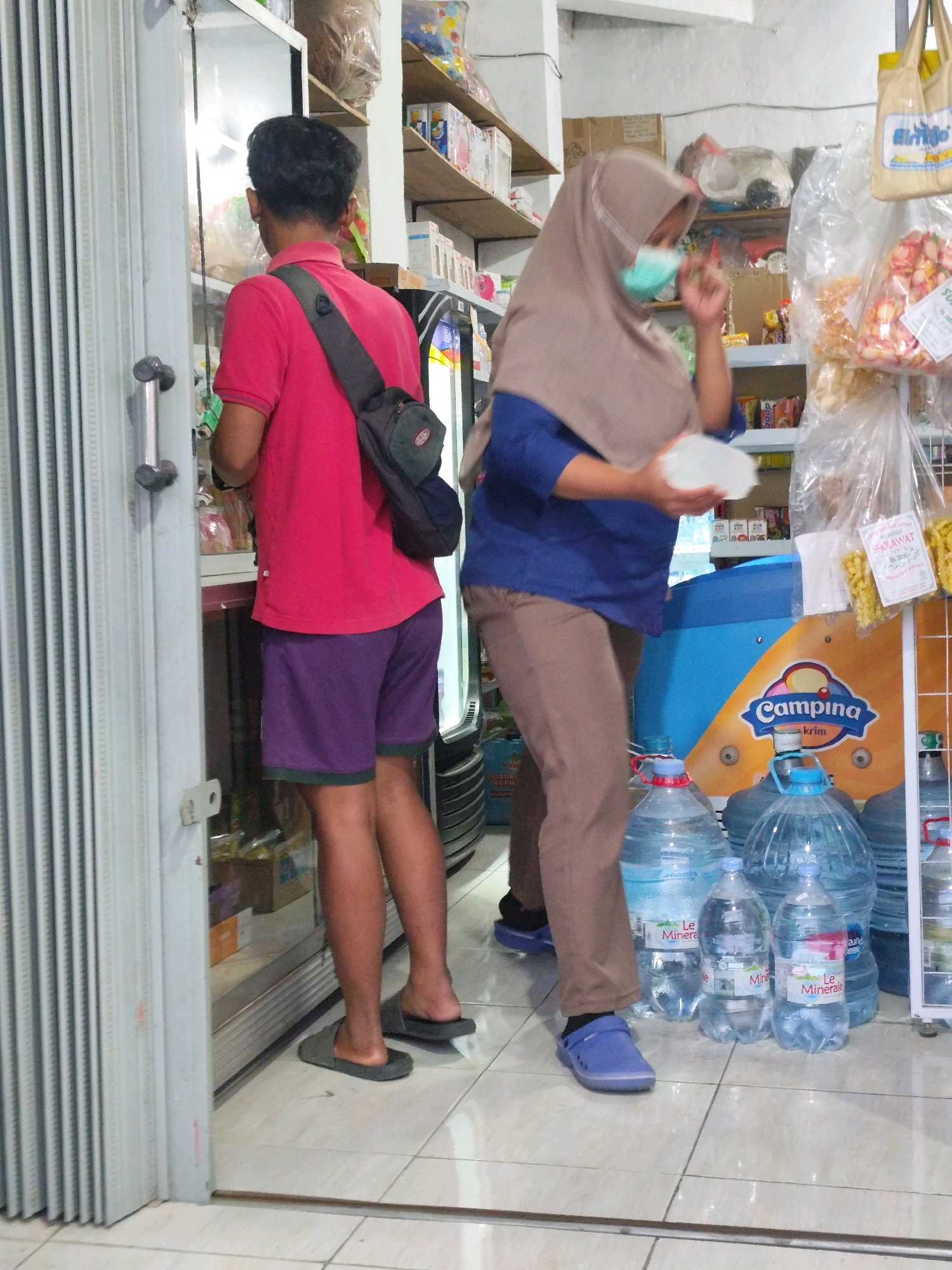The image captures an indoor scene at what appears to be a small convenience store or warung, located in Bambang Lipuro, Indonesia. In the foreground, a person wearing a pink t-shirt, dark shorts, and a black sling bag over their shoulder stands with their back to the camera, facing a refrigerator or display case. To the right, another individual is visible from the chest down, wearing a beige hijab, a blue shirt, and brown pants. This person is holding a white plastic lid or container. In front of them are several large blue-gallon water bottles and smaller plastic water bottles, some bearing labels. Shelves stocked with various products are visible in the background, along with hanging bags of snacks. The floor is tiled. The overall lighting suggests an indoor commercial setting.