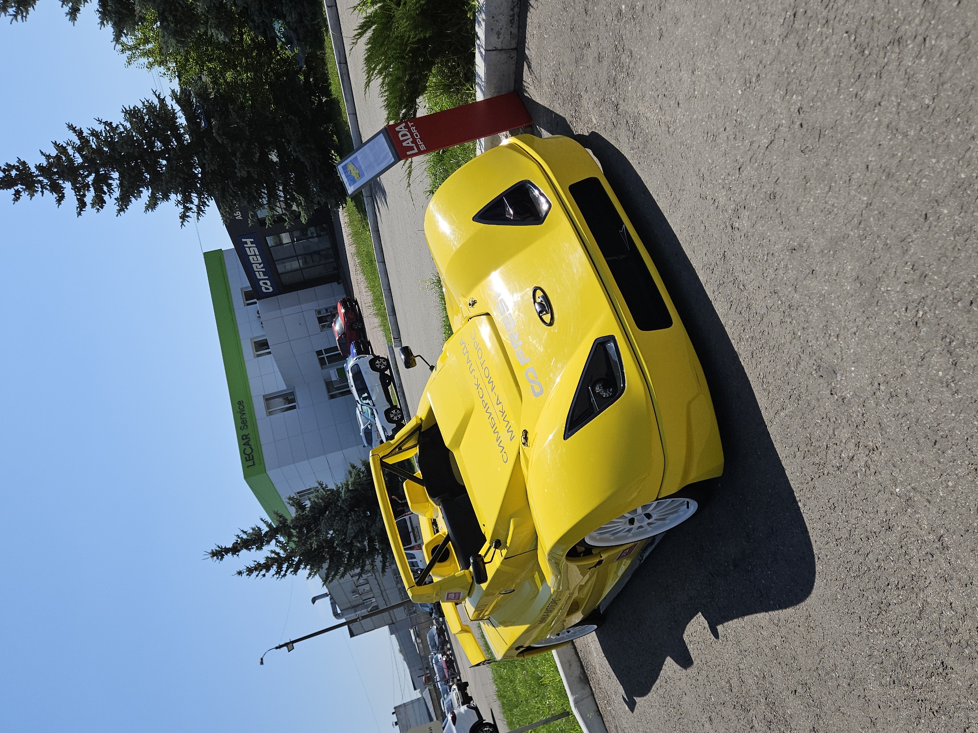 The image captures a yellow sports car parked on an asphalt surface in front of a modern building complex. The building on the left has a sign that reads "LECAR Service," and the building on the right has a sign that reads "GO FRESH." In the background, there are other vehicles and trees. A red signpost with "LADA SPORT" branding is also visible to the right of the car. The scene appears to be an outdoor area of a car dealership or service center, likely in the city of Ulyanovsk, Russia, given the "LADA SPORT" branding which is a Russian automotive brand. The weather is clear and sunny, as indicated by the bright blue sky and the shadows cast on the ground. There are no people visible in the image.