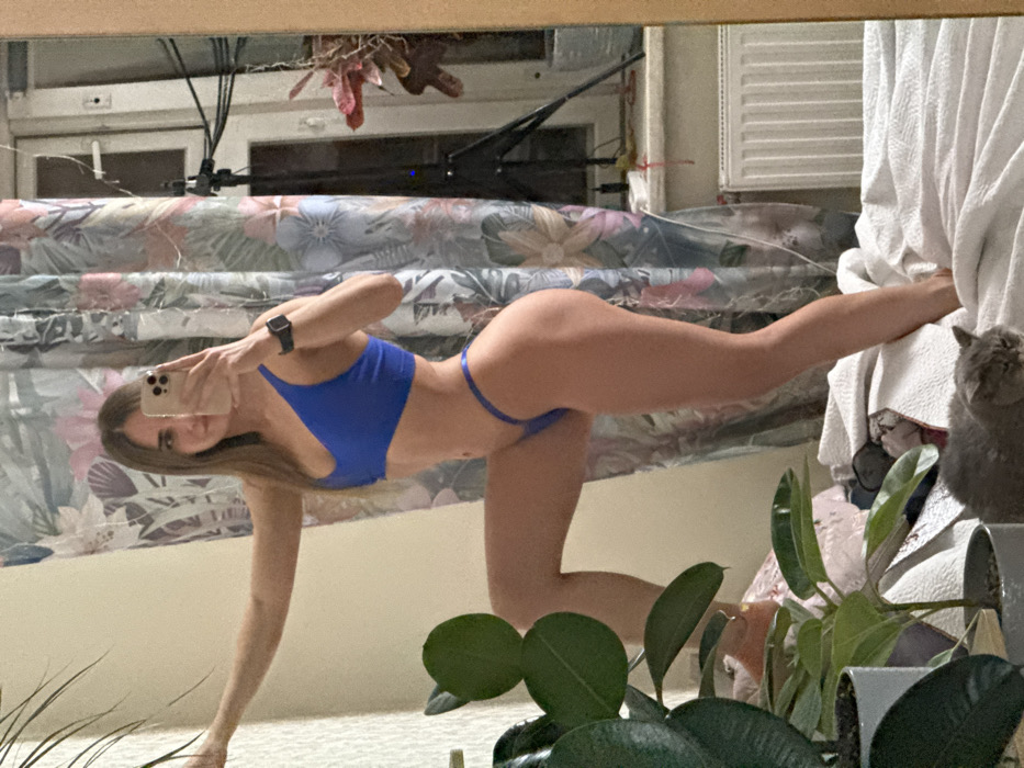 A woman in a blue bikini strikes a pose in front of a mirror, holding a smartphone up to take a selfie. She's on her hands and knees, arching her back. A patterned headboard is behind her. To her right, a white curtain is partially visible, with a gray cat perched on it. A houseplant with large green leaves partially obscures the view in the foreground.
