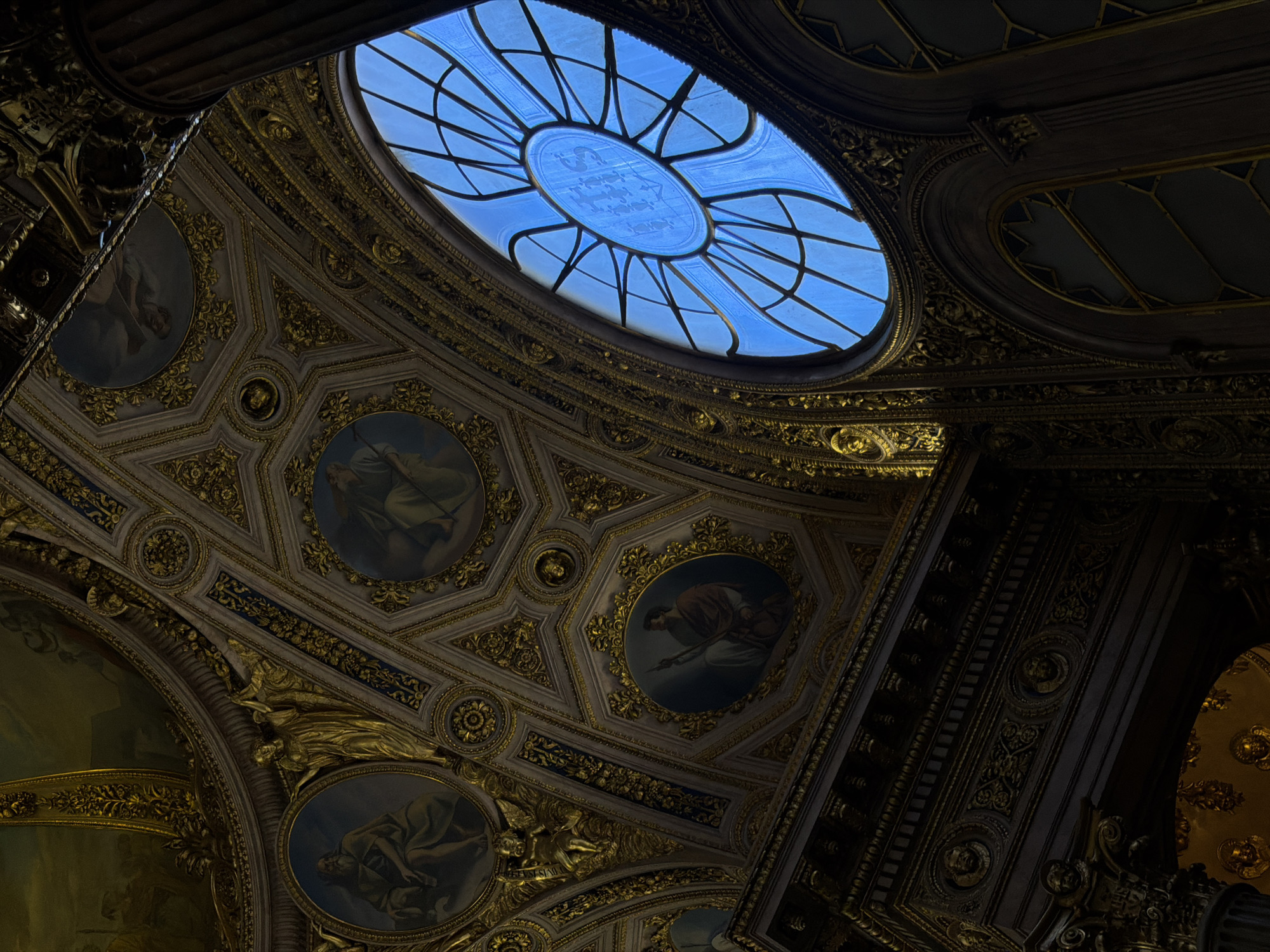 The image captures an ornate interior, likely a historic building or a museum, with a focus on the richly decorated ceiling and a large, blue-tinted arched window. The ceiling is a complex mosaic of hexagonal panels adorned with intricate gold detailing, possibly gilded plasterwork or stucco. Several circular paintings, depicting figures in classical attire, are embedded within the ceiling's design. These paintings are set within gold frames that echo the geometric patterns of the ceiling.
To the right of the frame, a large, elliptical window with a radial pattern of leaded glass dominates the upper portion. The glass is a vibrant blue, suggesting it is either stained glass or illuminated by a bright blue sky. Below this, a series of arched windows, also with geometric patterns in their panes, are visible.
The lower part of the image shows a dark, richly carved wooden element, possibly part of an entablature or a grand archway. This section features detailed reliefs, including what appear to be faces or masks and floral motifs, all rendered in dark wood with hints of gold. A large, fluted column, also dark and substantial, is visible on the far right edge of the image, adding to the sense of grandeur and architectural scale.
The lighting in the scene is dramatic, with strong contrasts between the illuminated window and the shadowed interior. This creates a sense of depth and highlights the textures and details of the ornamentation.
While the prompt mentions Mozzo, Italy, there are no discernible visual cues in the image that specifically place it in that location. The architectural style is reminiscent of European classical or baroque interiors, but without further context, a precise identification of the building or city is not possible from the image alone.
