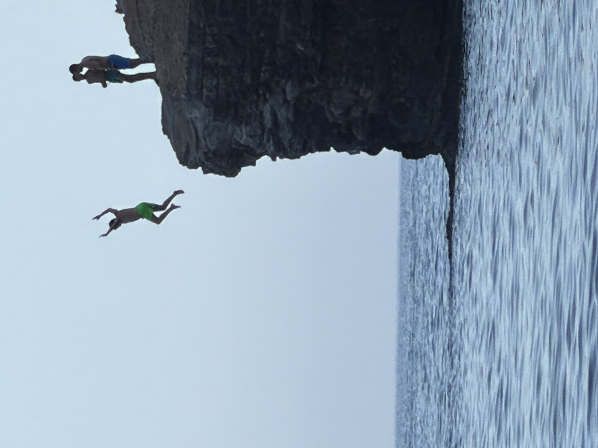 In Budva, Montenegro, three young men are captured in a moment of daring. One individual is mid-air, having just leaped from a rocky cliff into the calm, blue sea. His body is suspended horizontally, arms outstretched, mid-jump. Above and behind him, two other men stand on the edge of the cliff, observing. The cliff face is rugged and dark, contrasting with the pale sky and the shimmering water. The scene evokes a sense of adventure and freedom, set against the natural beauty of the Montenegrin coast.