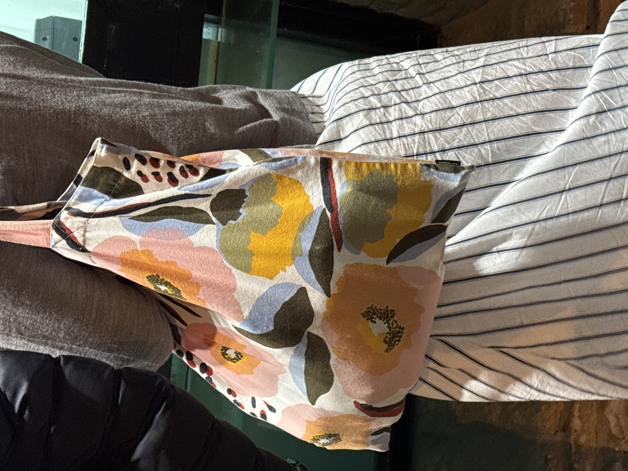 The image shows a close-up of textiles. From left to right, there is a dark, puffy fabric (possibly a jacket), a gray cloth, a floral-patterned bag, and a white striped pillowcase. The objects are stacked and rumpled. The setting is unclear, but the background features elements that look like a stone or concrete wall. The location is unspecified, and there's no indication of Tbilisi, Georgia or people, actions, or interactions.