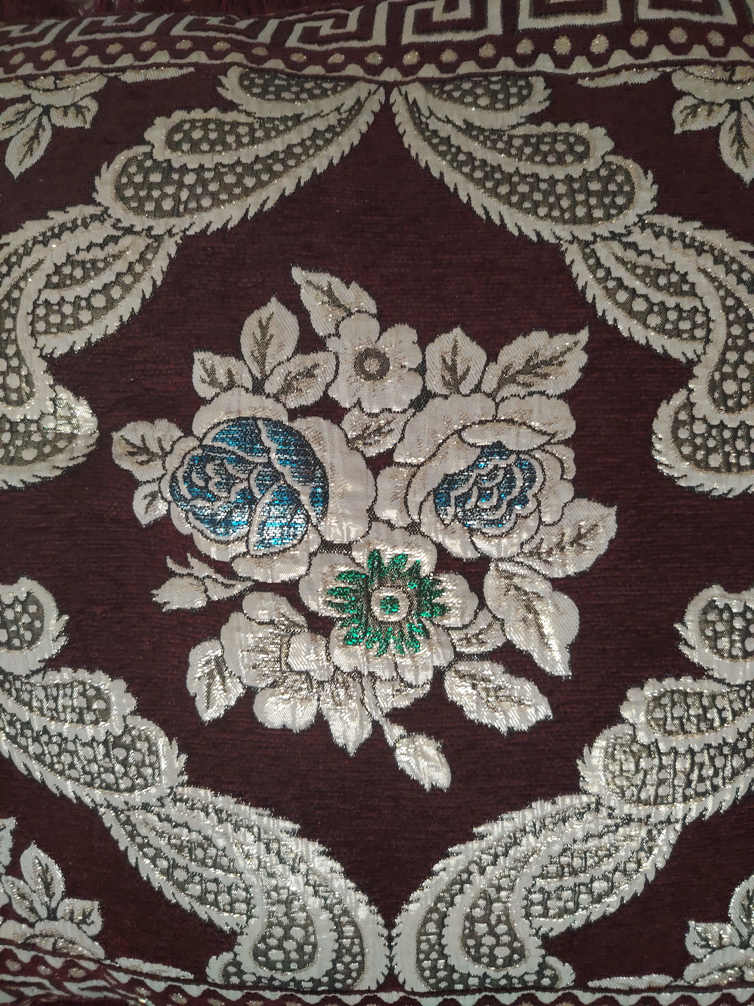 The image displays a close-up of a richly patterned fabric, likely a textile or upholstery. The dominant color is a deep maroon, providing a backdrop for intricate metallic embroidery. The embroidery features stylized floral motifs and paisley-like shapes in silver and gold threads, with accents of bright blue and green in the center of some flowers. The texture of the fabric appears to be woven, with a slight sheen from the metallic threads. The pattern is dense and elaborate, suggesting it is part of a decorative item such as a cushion, tapestry, or garment. There are no people or any indication of a specific location within the city of Pitnak, Uzbekistan. The image solely focuses on the detailed textile design.