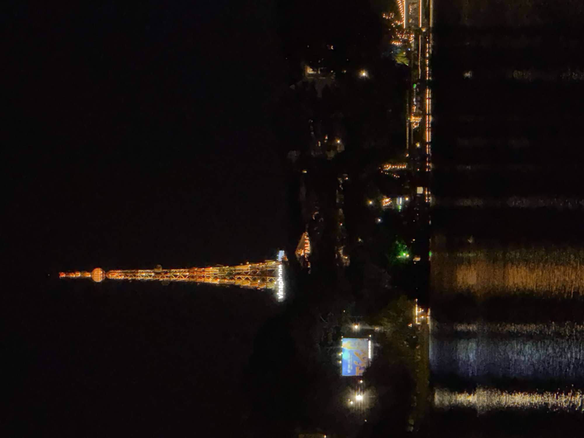 The image shows a night view of the city of Da Lat, Vietnam, featuring a tall, illuminated radio tower as the central subject. The tower, likely VinaPhone, is lit with orange and yellow lights, creating a prominent structure against the dark night sky. Below the tower, a cluster of buildings and lights are visible, suggesting a developed area. To the left of the tower, a large illuminated billboard with blue and yellow graphics is present. The foreground is dominated by a body of water, likely Xuan Huong Lake, with reflections of the lights from the city and the tower shimmering on its surface. The overall scene depicts a vibrant urban landscape at night, with the illuminated tower serving as a focal point. There are no discernible people or specific actions captured in the image.