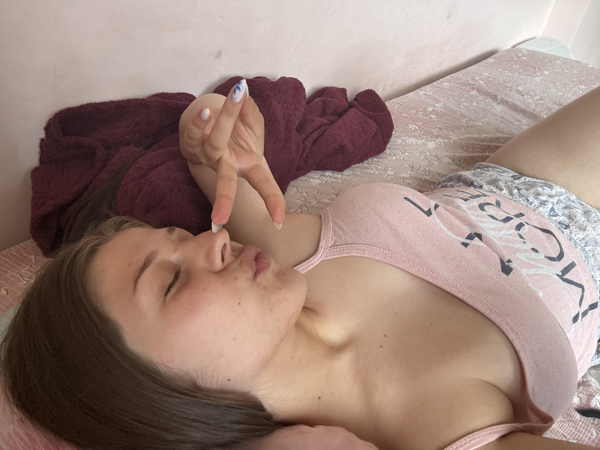 This is a medium shot, close-up, indoor photograph of a young woman lying on her side on a bed. The woman has long brown hair and is wearing a pink tank top and patterned shorts. Her eyes are closed and her lips are puckered. She is holding up her left hand in a peace sign gesture. The background consists of a white wall and a bed with a patterned pink and white comforter. A dark red blanket is draped over the bed. The lighting suggests it is daytime, possibly morning or afternoon. The overall mood is relaxed and playful. There is visible text on the woman's tank top, but it is partially obscured and difficult to read. The nails on the woman's hands are painted with white and blue designs.