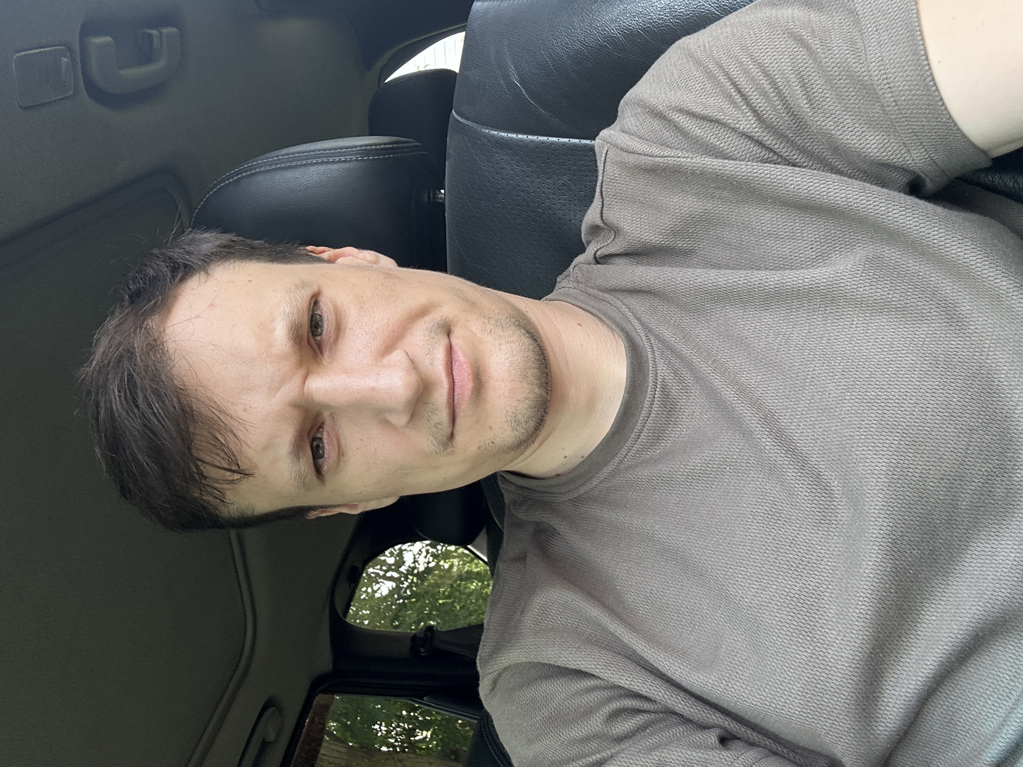 The image shows a selfie of a man sitting in the driver's seat of a car. He is looking directly at the camera with a slightly furrowed brow. The man is wearing a brown t-shirt. The interior of the car, including black leather seats and a sunroof, is visible. Outside the car window, trees and foliage can be seen, suggesting a suburban or rural setting. The image does not provide any information that would allow for the determination of its location in Makiv, Ukraine.