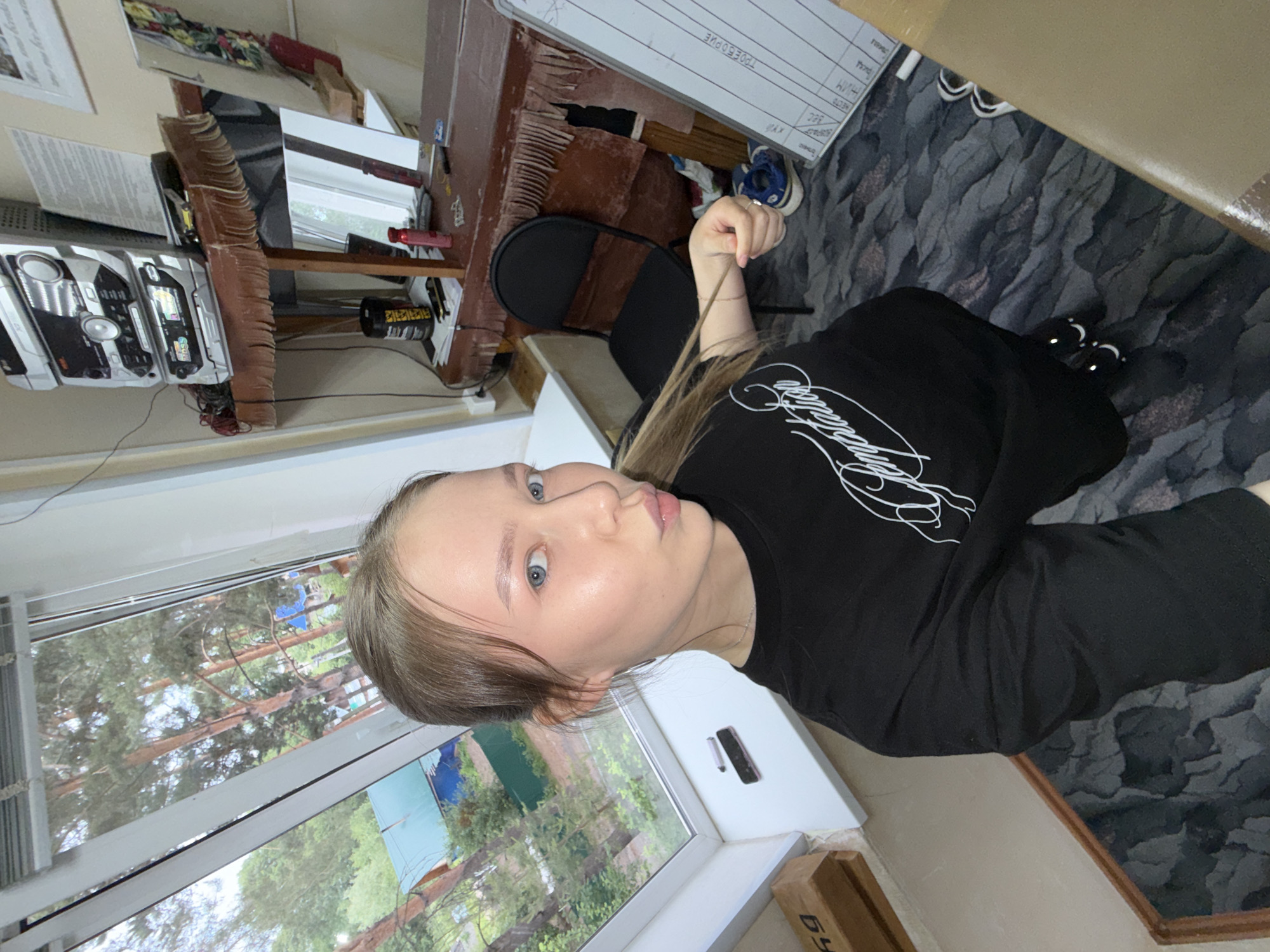 The image shows a young woman with blonde hair and blue eyes taking a selfie in an indoor setting. She is wearing a black t-shirt with white text that reads "Temptation" and black pants or leggings. Her lips are pursed, and she is holding a strand of her hair.
The environment appears to be a room with a large window that looks out onto trees and a green roof. Inside the room, there is a desk with various items on it, including a stereo system, papers, and a water bottle. A black chair is positioned in front of the desk, and a whiteboard with writing on it is leaning against the wall. The floor is covered with a patterned carpet. The overall impression is of a casual, perhaps home or office, environment.
Given the details provided in the prompt, this location is identified as being in the city of Chaikovsky, Russia.